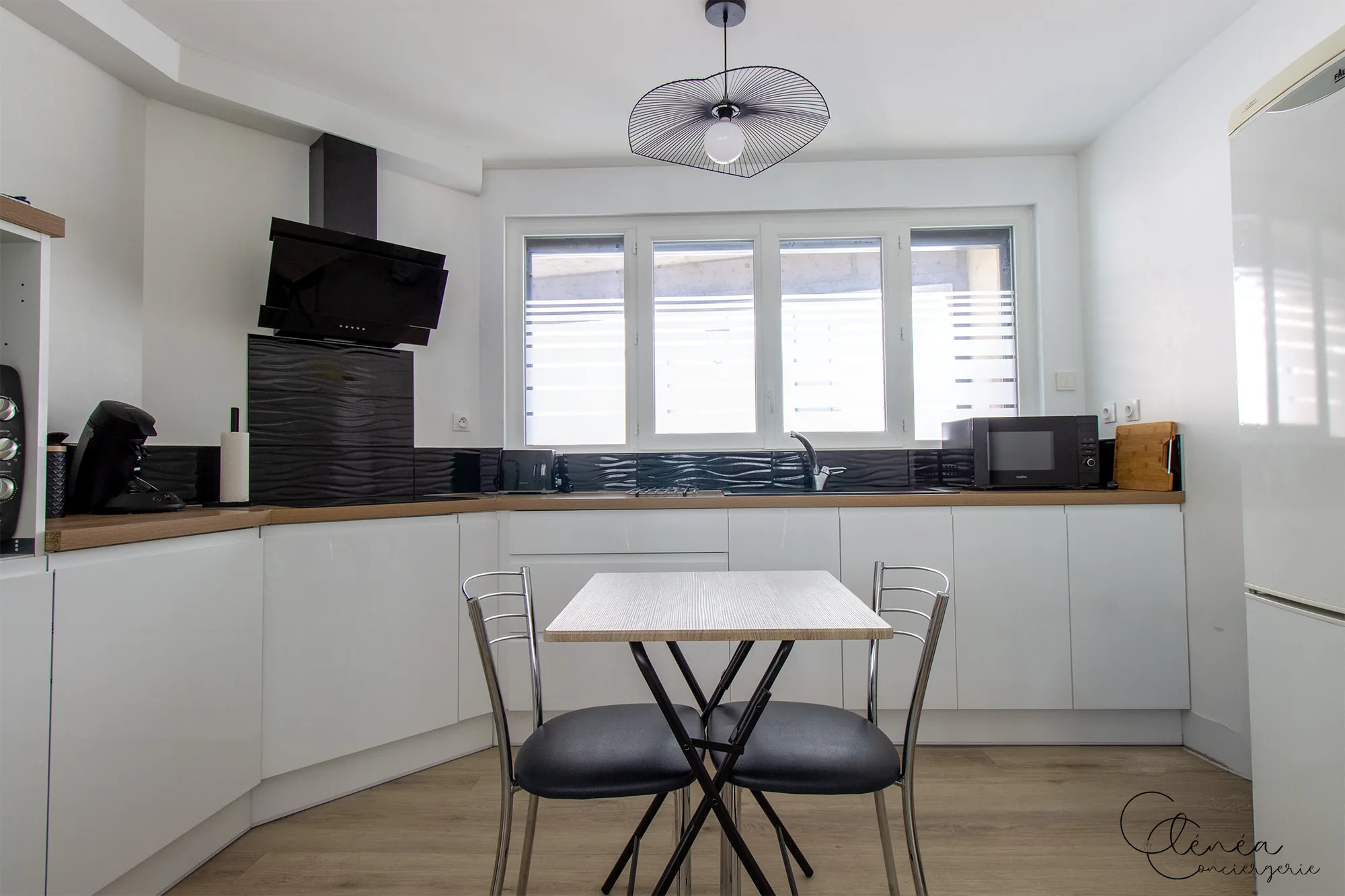 Au Cœur des Lices — Vue d'ensemble de la cuisine : plan de travail en angle, table repas pour deux et réfrigérateur.