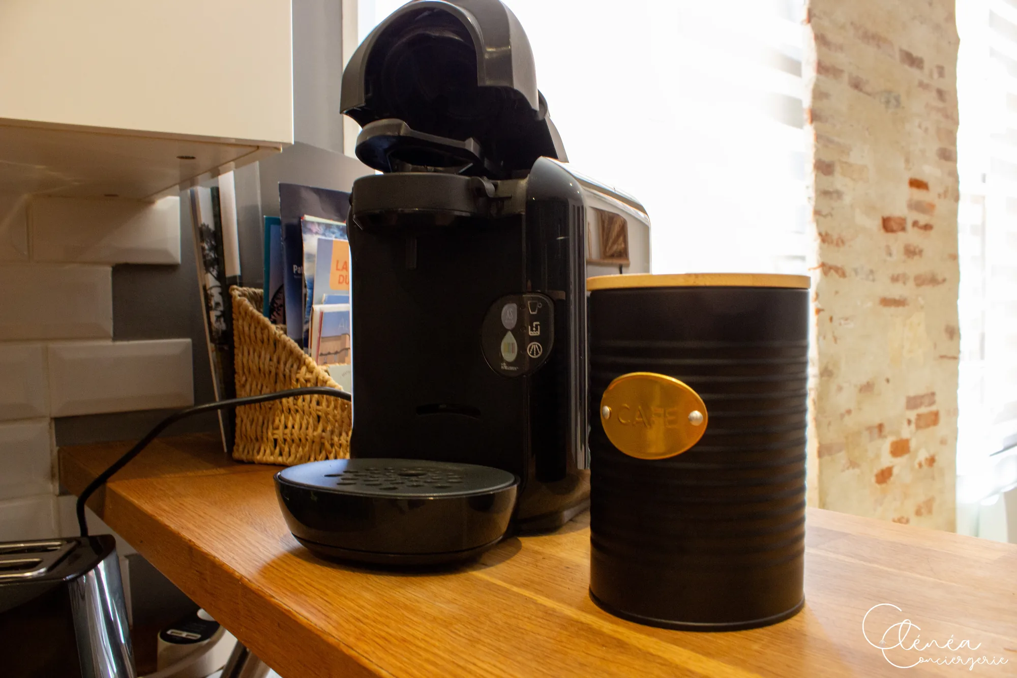 Chez Ludi et Pierre — Machine à café à capsules et boîte à café posées sur le plan de travail en bois, devant un mur de briques