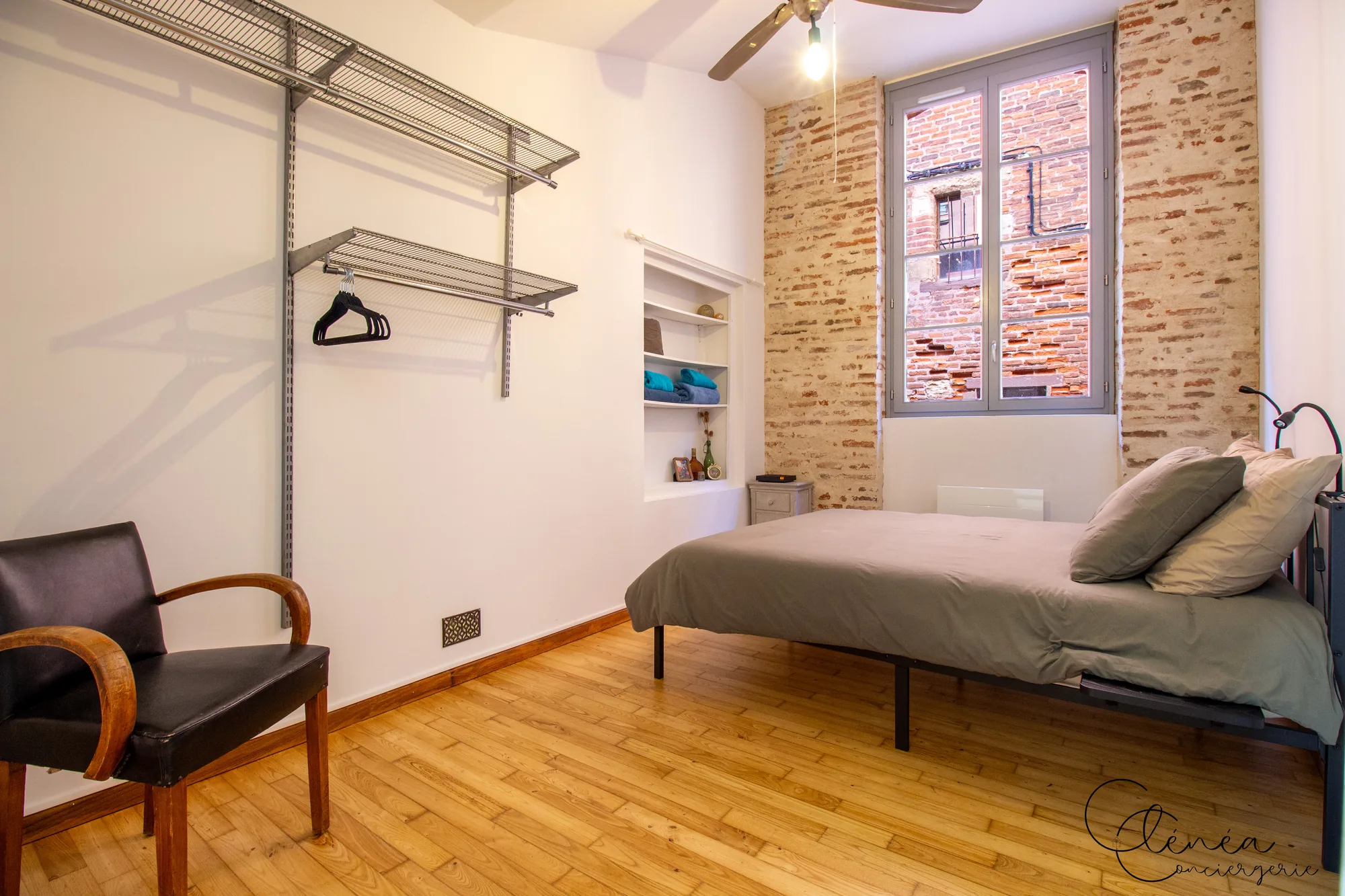 Chez Ludi et Pierre — Chambre 1 avec ventilateur de plafond, penderie ouverte, niche de rangement et fenêtre donnant sur un mur de briques