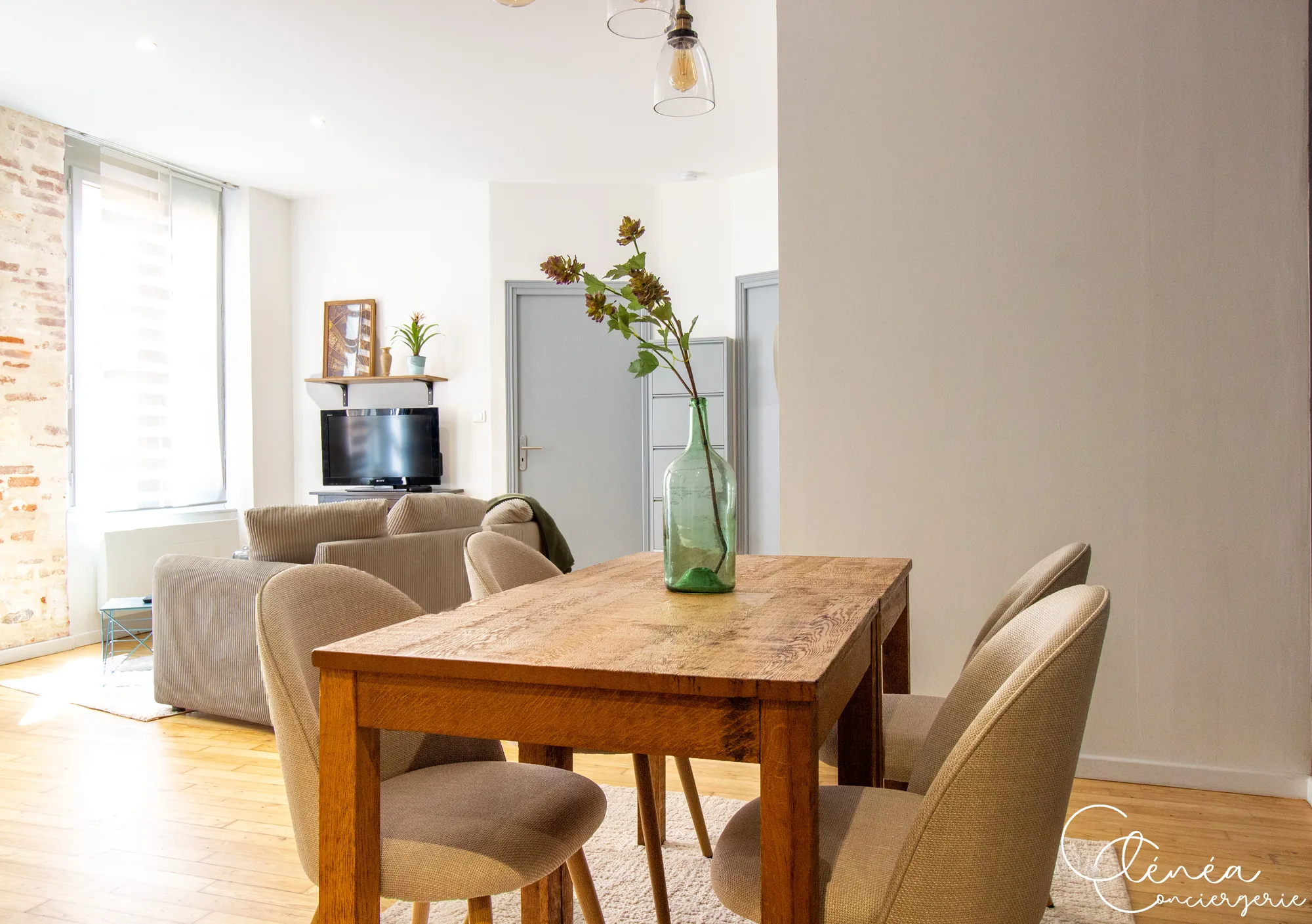Chez Ludi et Pierre — Table à manger en bois et ses chaises, avec le canapé, la télévision et un mur de briques en arrière-plan