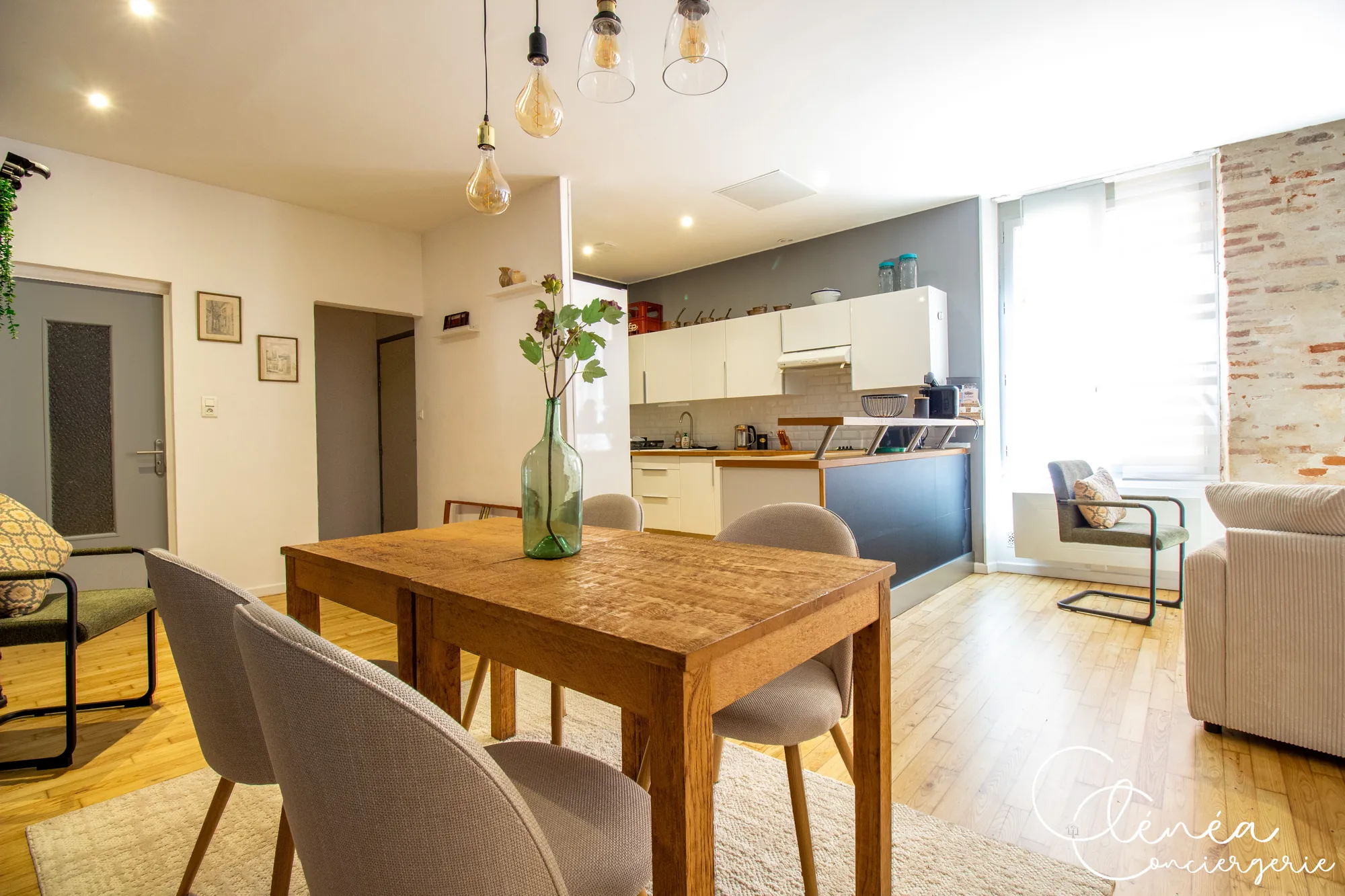 Chez Ludi et Pierre — Pièce de vie avec table à manger, canapé et cuisine ouverte séparée par un bar, éclairée par de hautes fenêtres