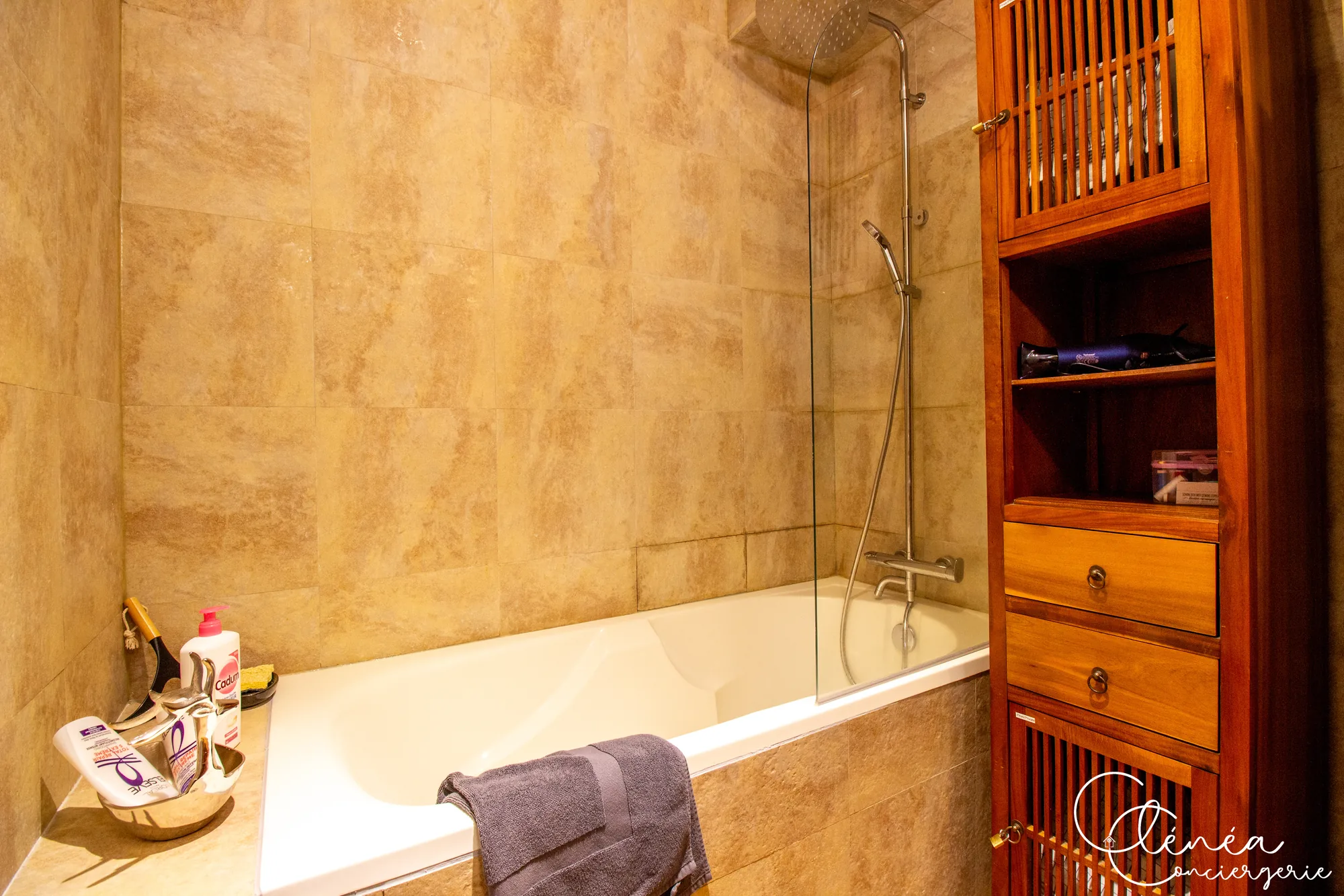 Chez Ludi et Pierre — Baignoire avec douchette et paroi vitrée, à côté d’une colonne de rangement en bois