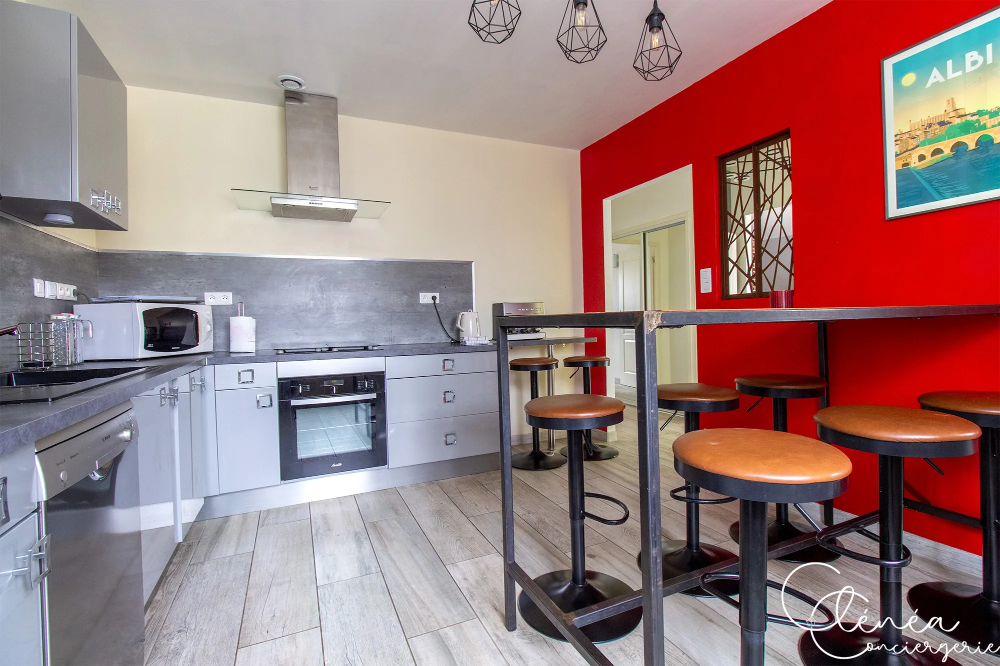 L’Escapade Albigeoise — Cuisine avec four, hotte et mur rouge orné d’une affiche d’Albi, devant la table mange-debout et ses tabourets