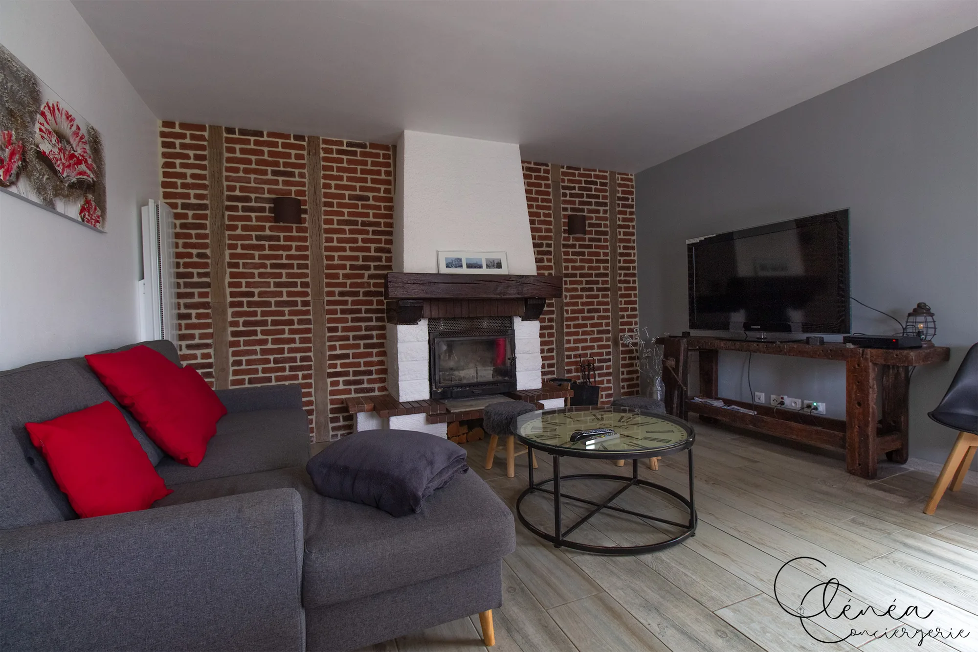 L’Escapade Albigeoise — Salon avec canapé gris à coussins rouges, cheminée à foyer fermé sous linteau en bois et télévision