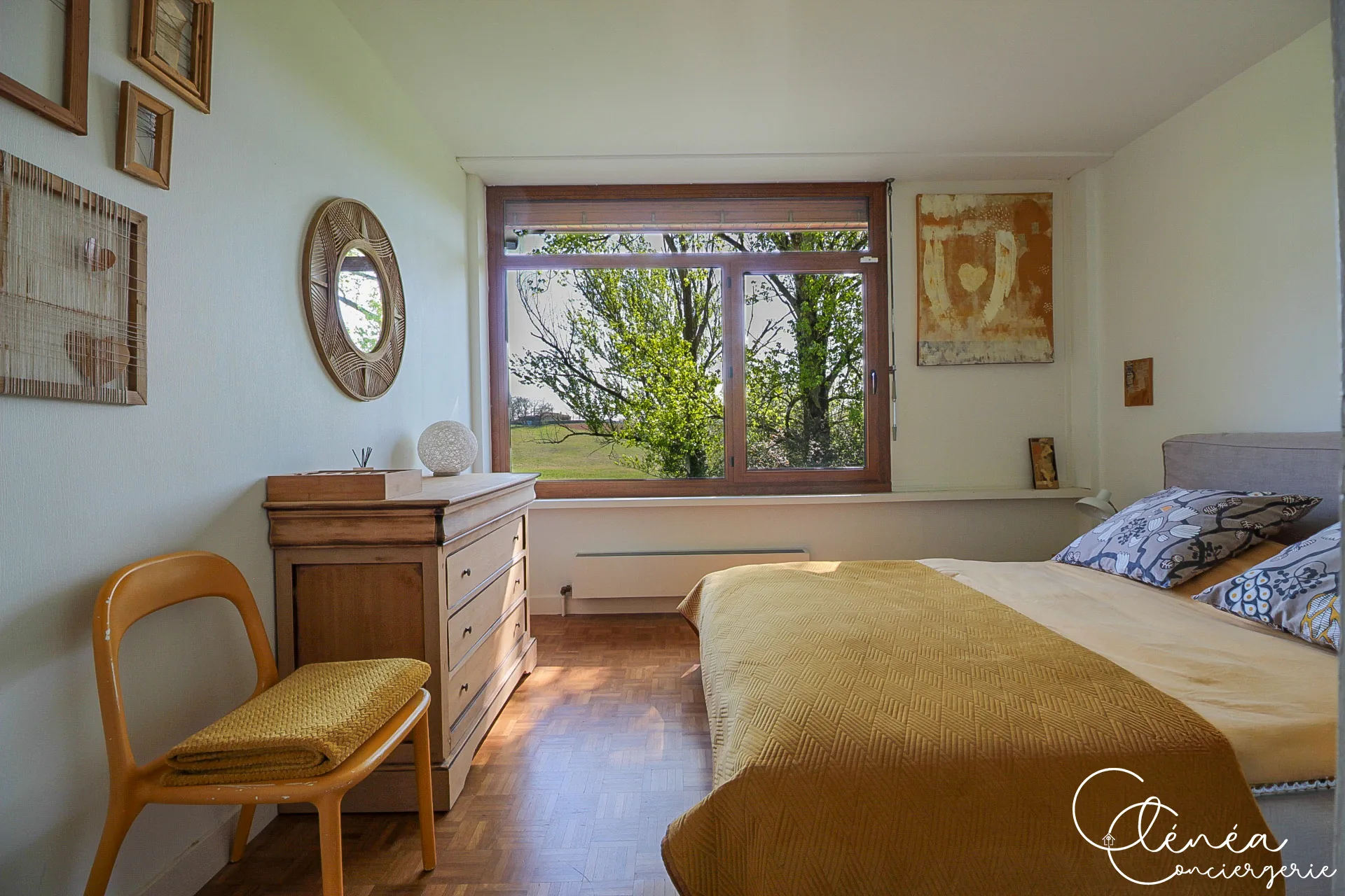 GreenOak — Chambre avec lit double aux teintes ocre, commode en bois clair et chaise, fenêtre sur les arbres