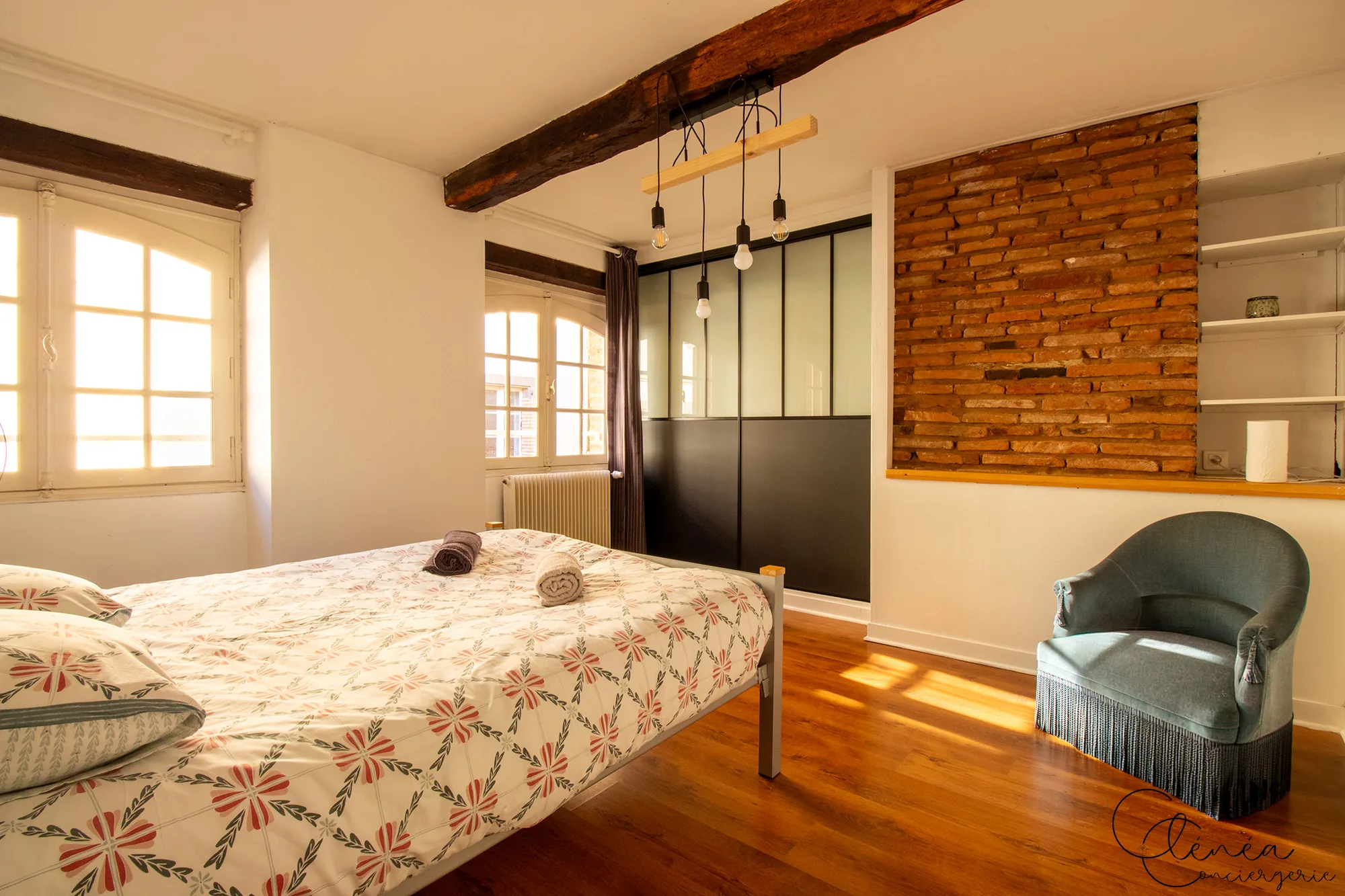 Le Quinze — Chambre avec lit double, mur de briques apparentes, verrière d’atelier et fauteuil crapaud bleu