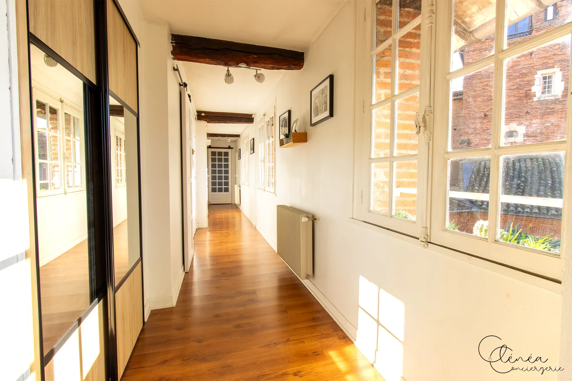 Le Quinze — Long couloir en parquet bordé de fenêtres, avec placard à portes miroir et cadres accrochés au mur
