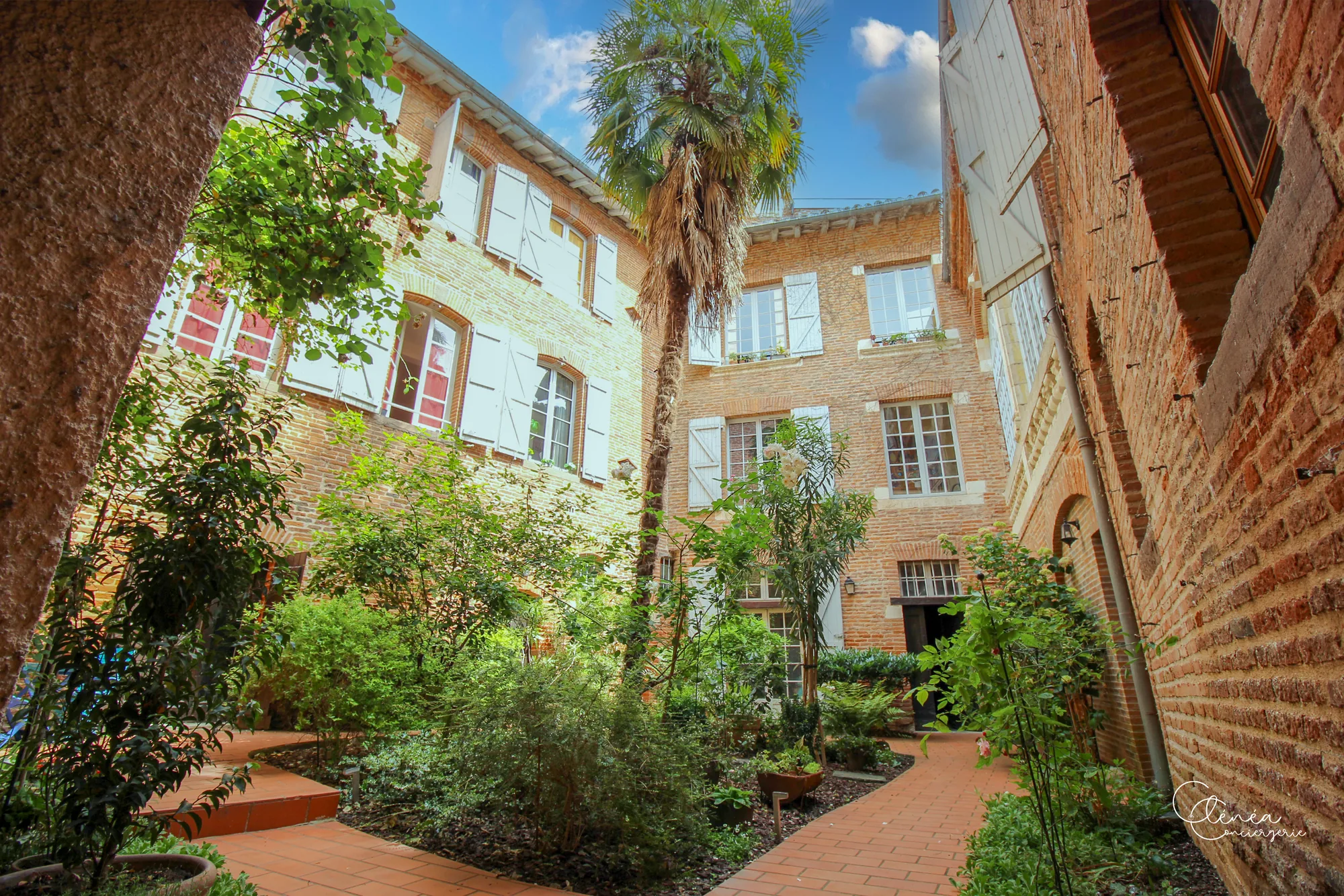 Le Quinze — Cour intérieure arborée bordée de façades en brique rouge, avec allée pavée et palmier