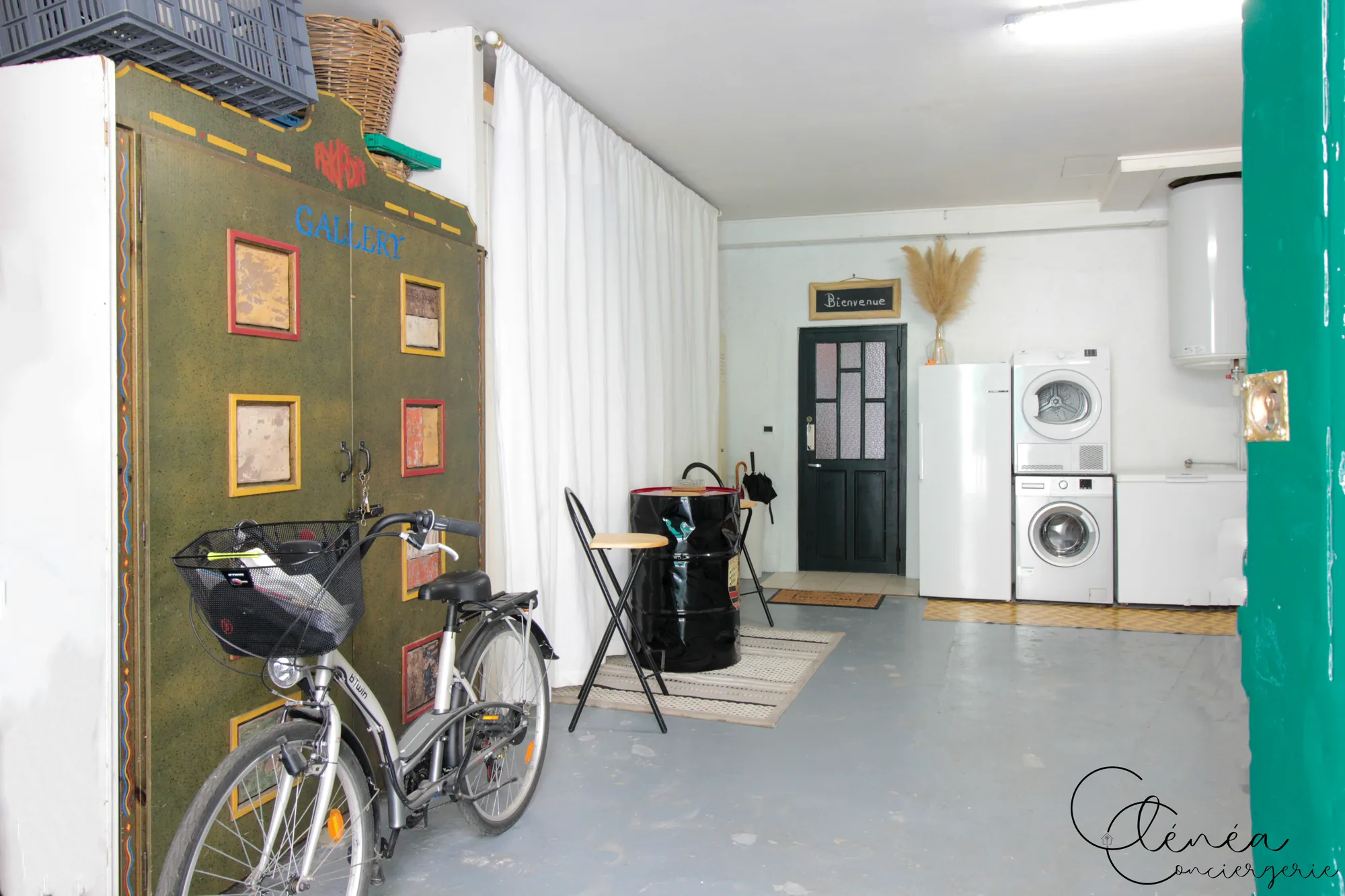 Saudade d’Albi — Garage privatif avec un vélo appuyé contre une porte en bois peinte, table haute, machine à laver et sèche-linge au fond près de la porte d’entrée du logement.