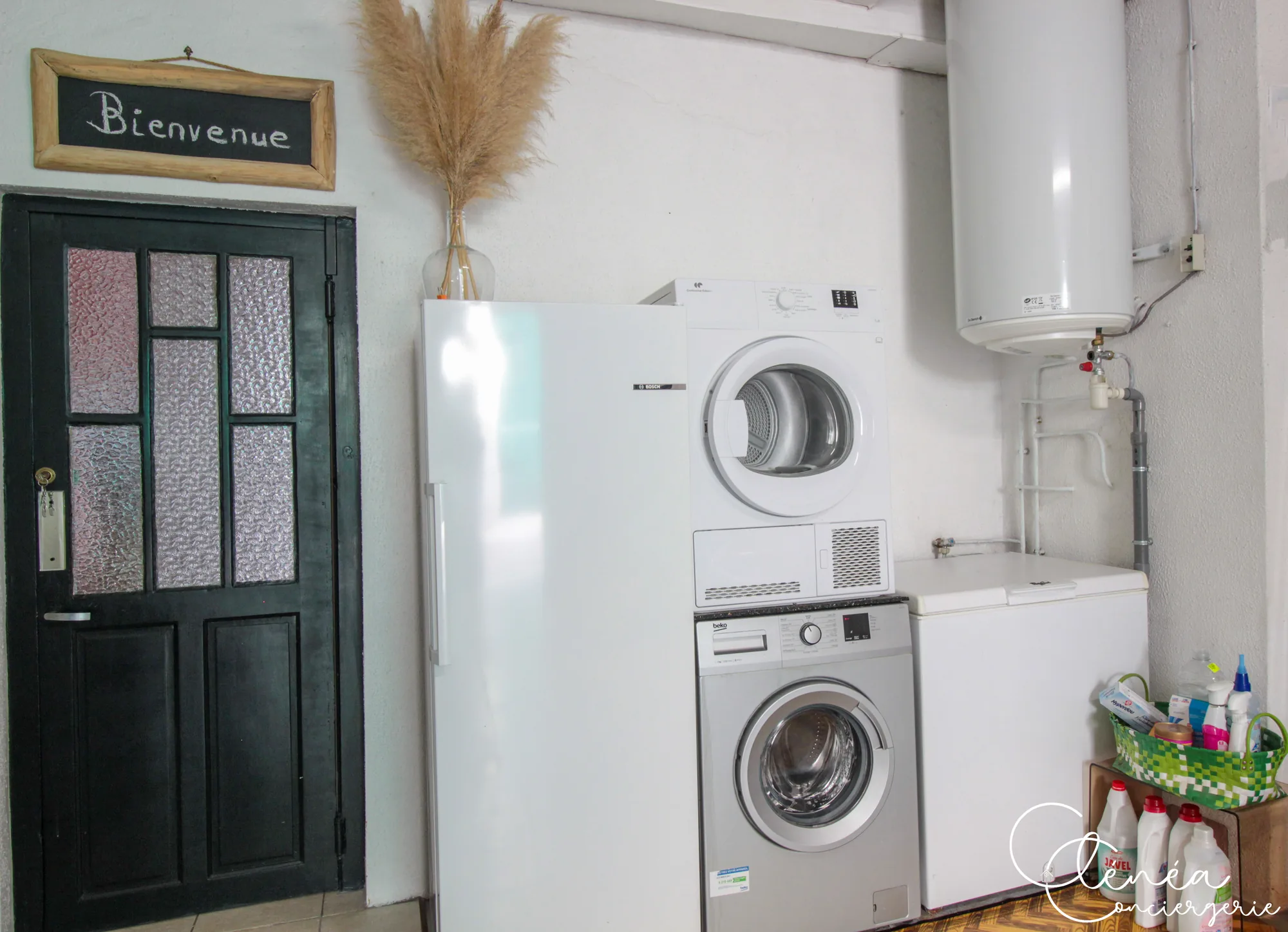 Saudade d’Albi — Espace buanderie avec lave-linge surmonté d’un sèche-linge, réfrigérateur, congélateur coffre et porte d’entrée verte surmontée d’une ardoise « Bienvenue ».