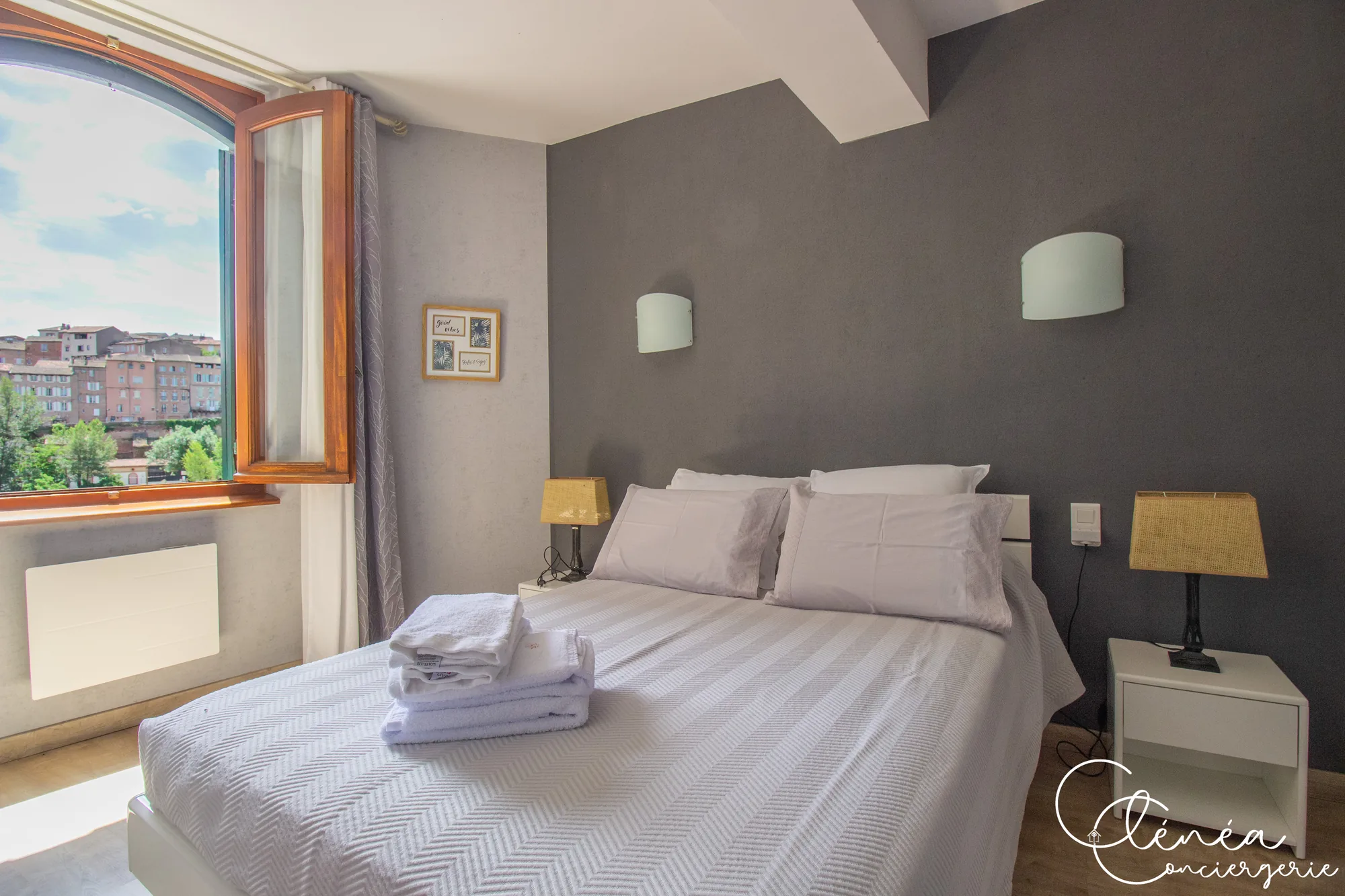 Saudade d’Albi — Lit double de la chambre devant une fenêtre en plein cintre ouverte sur les toits et les façades de brique d’Albi.