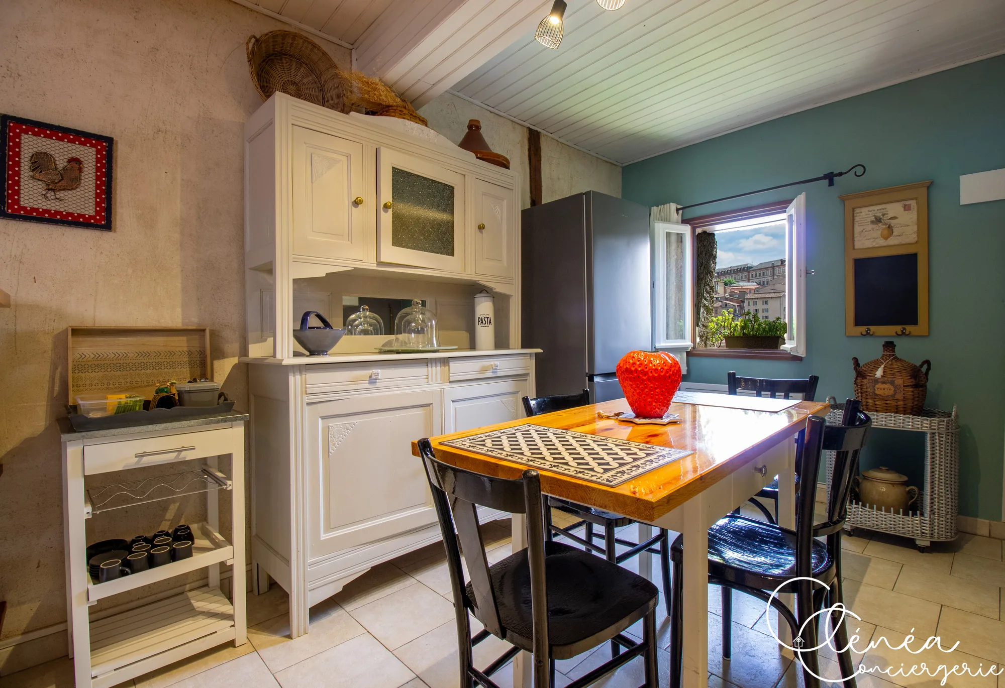 Saudade d’Albi — Coin repas de la cuisine avec buffet ancien blanc, réfrigérateur, table en bois et fenêtre ouverte sur les toits de la ville.