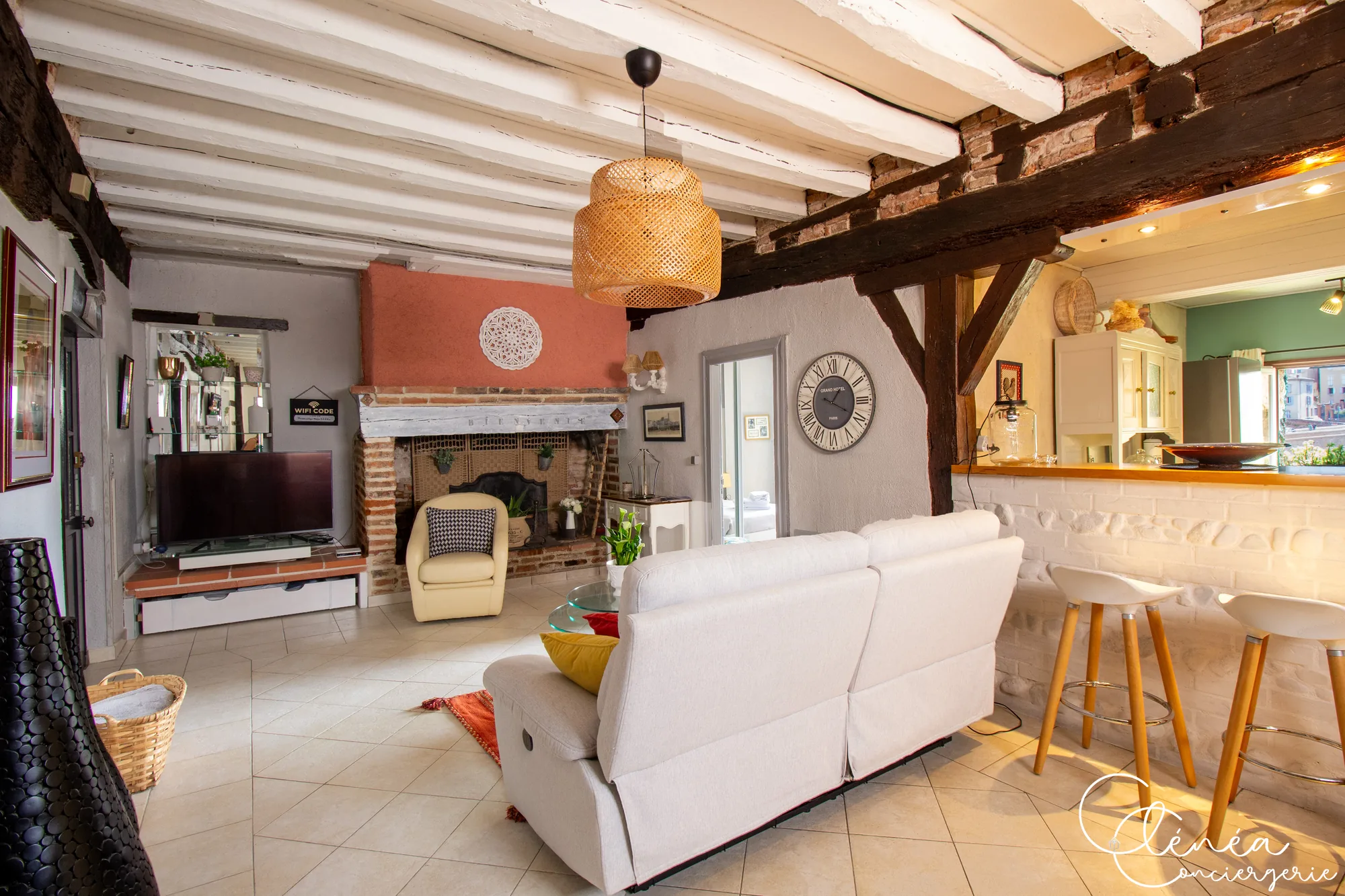 Saudade d’Albi — Pièce de vie sous poutres blanches avec cheminée en brique, télévision, canapé clair, fauteuil et bar avec tabourets côté cuisine.