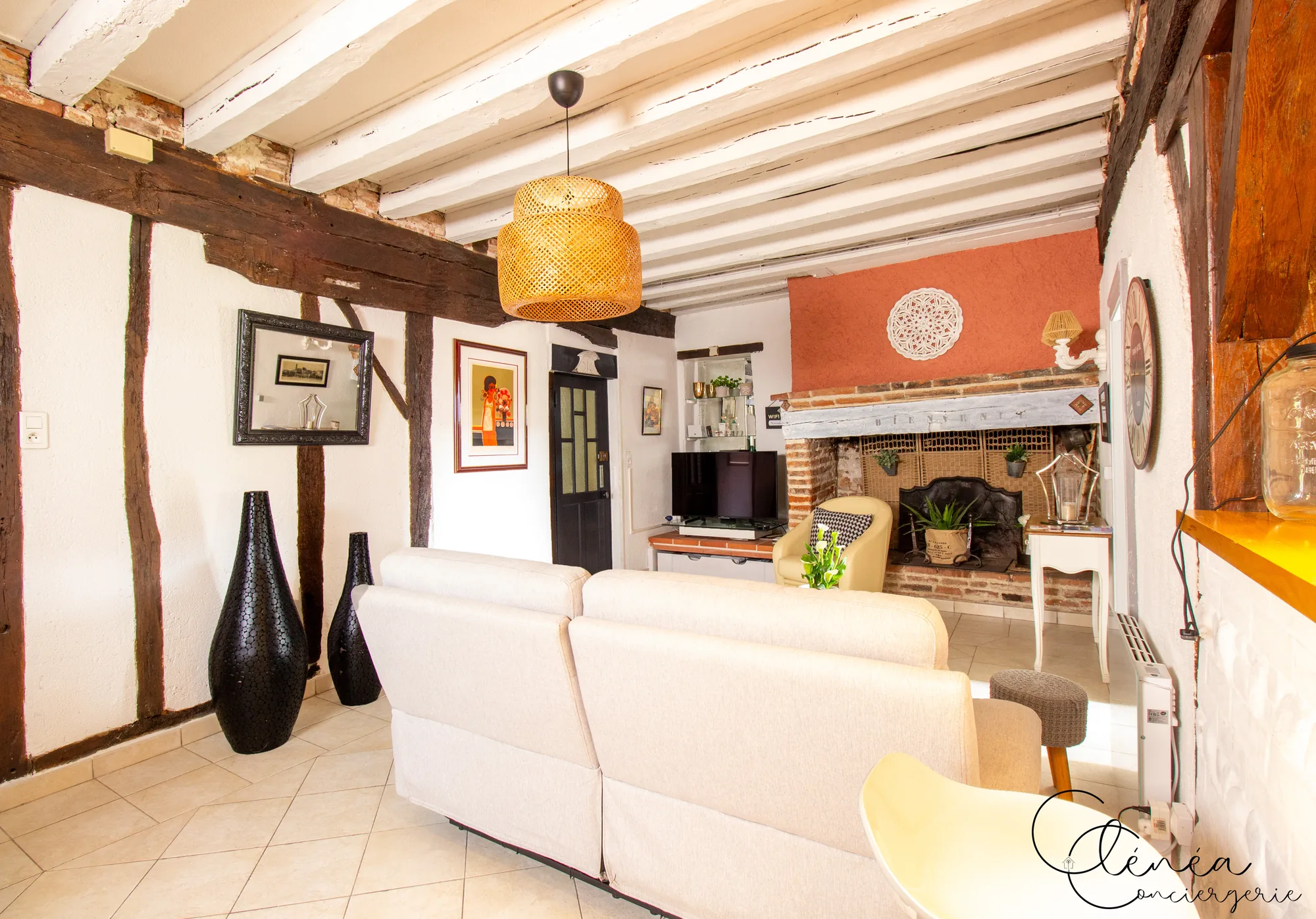 Saudade d’Albi — Salon vu depuis l’entrée, avec murs à colombages apparents, canapé clair, suspension en rotin et cheminée en brique au fond.