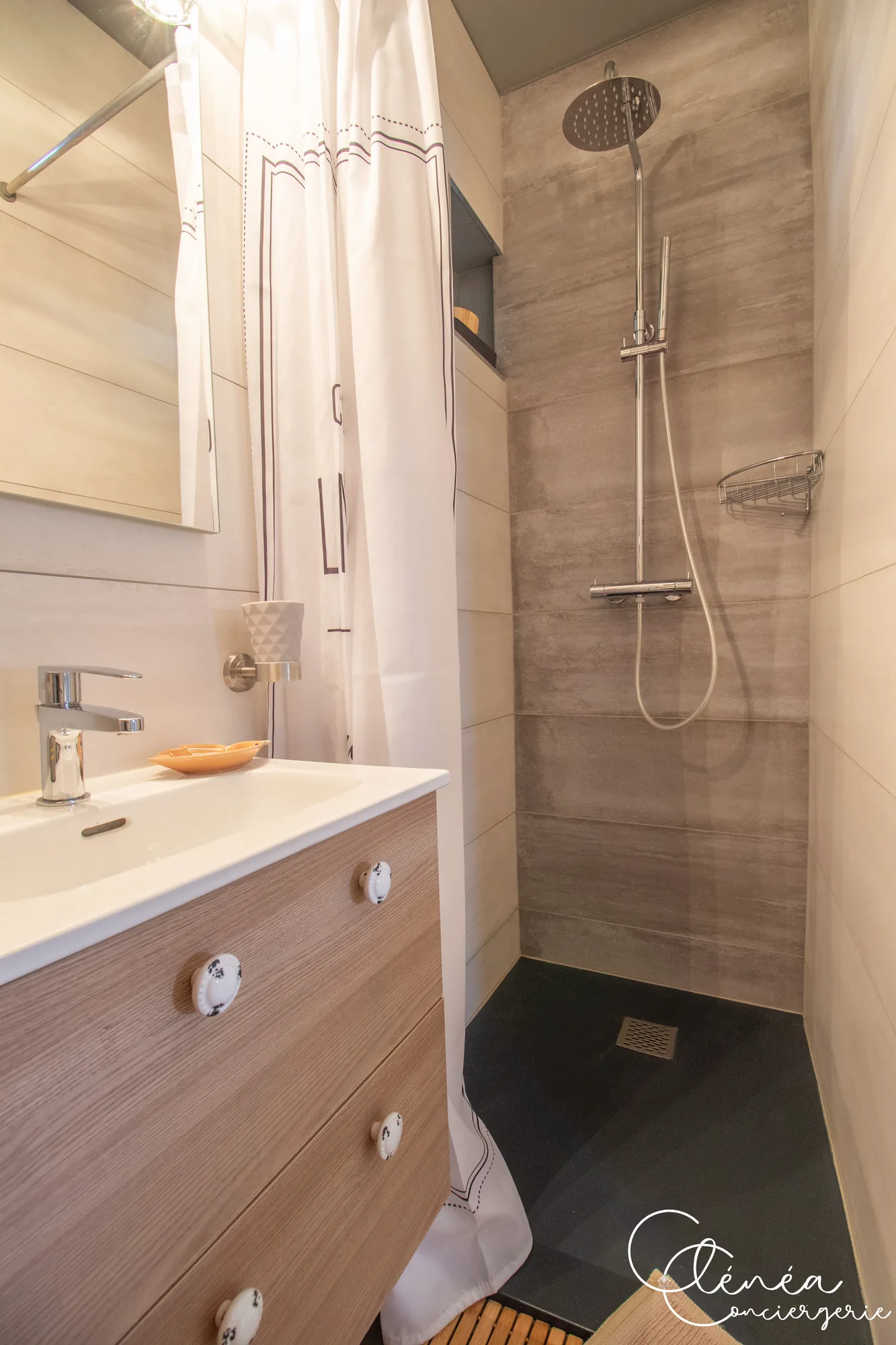 Saudade d’Albi — Salle de bain avec douche à l’italienne, colonne de douche et meuble vasque en bois clair surmonté d’un miroir.