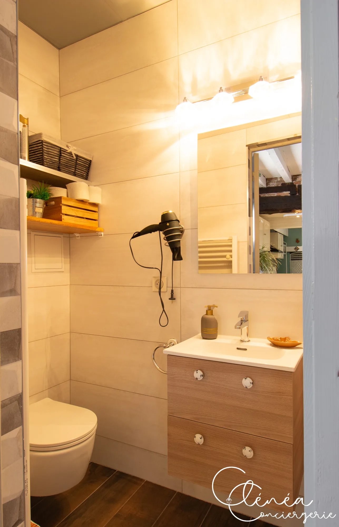 Saudade d’Albi — Salle de bain avec WC suspendu, meuble vasque en bois clair, miroir éclairé, sèche-cheveux mural et étagères de rangement.