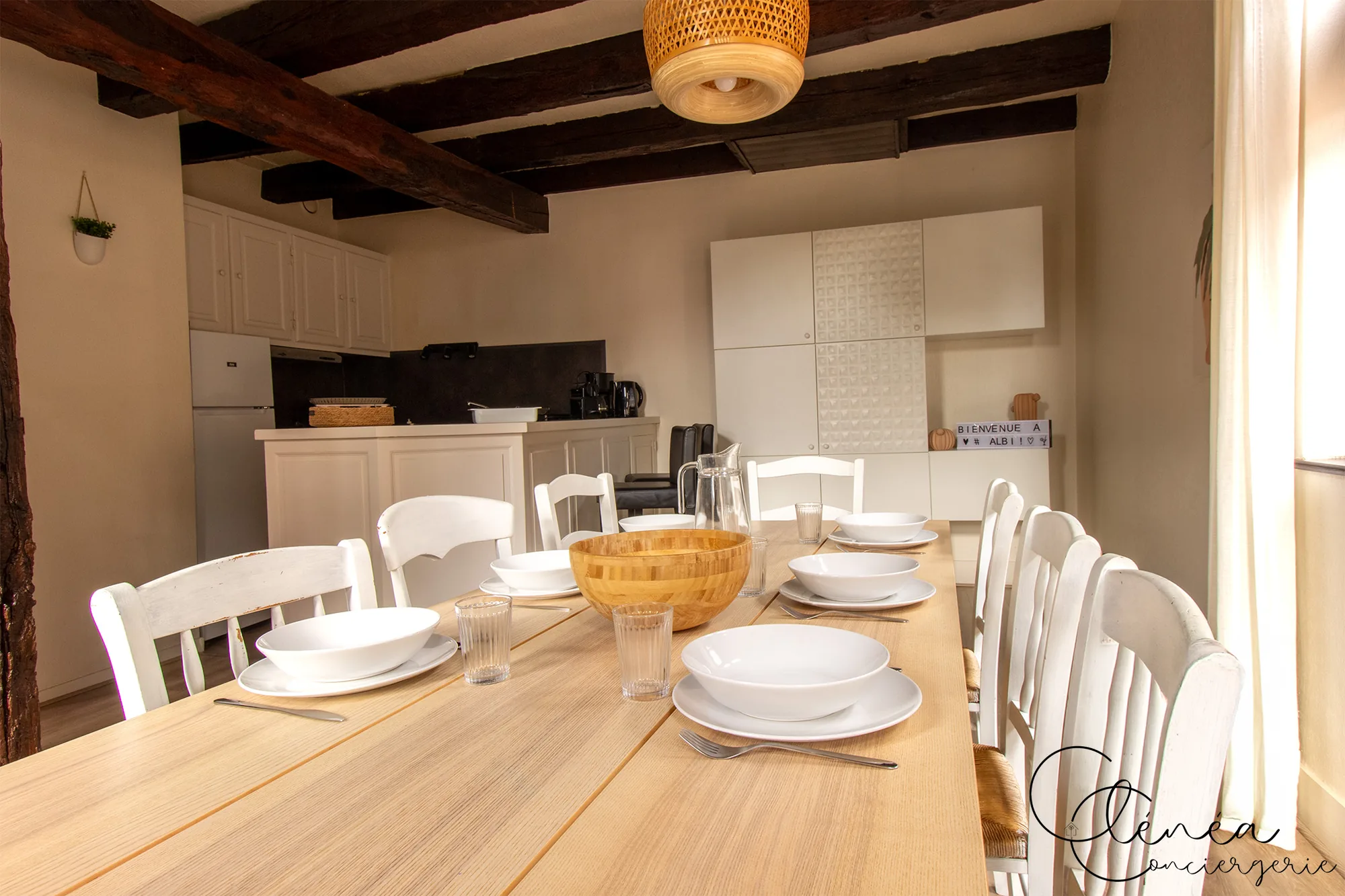 Sous les Toits de Sainte Cécile — Grande table en bois clair dressée pour plusieurs convives, chaises blanches et cuisine en arrière-plan sous les poutres