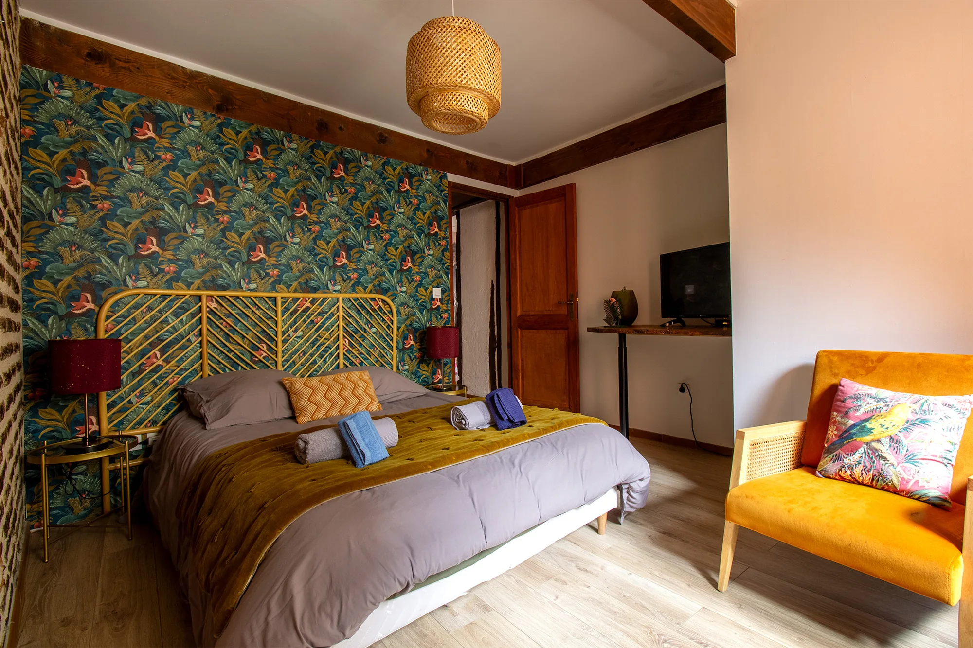 The Nest — Chambre avec lit double, tête de lit dorée devant un papier peint à motif végétal, télévision murale et fauteuil orange.