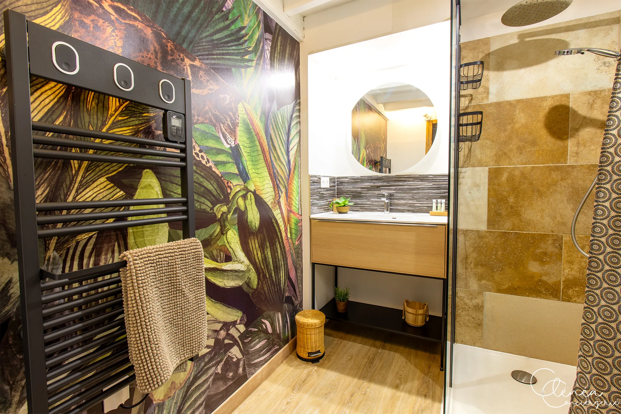The Nest — Salle de bain avec sèche-serviettes, papier peint tropical, meuble vasque surmonté d’un miroir rond et douche.