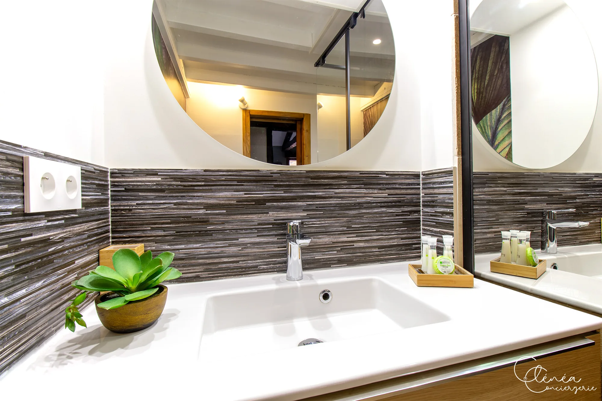The Nest — Vasque blanche, robinetterie chromée, miroir rond et petits flacons de produits de toilette sur un plateau en bois.