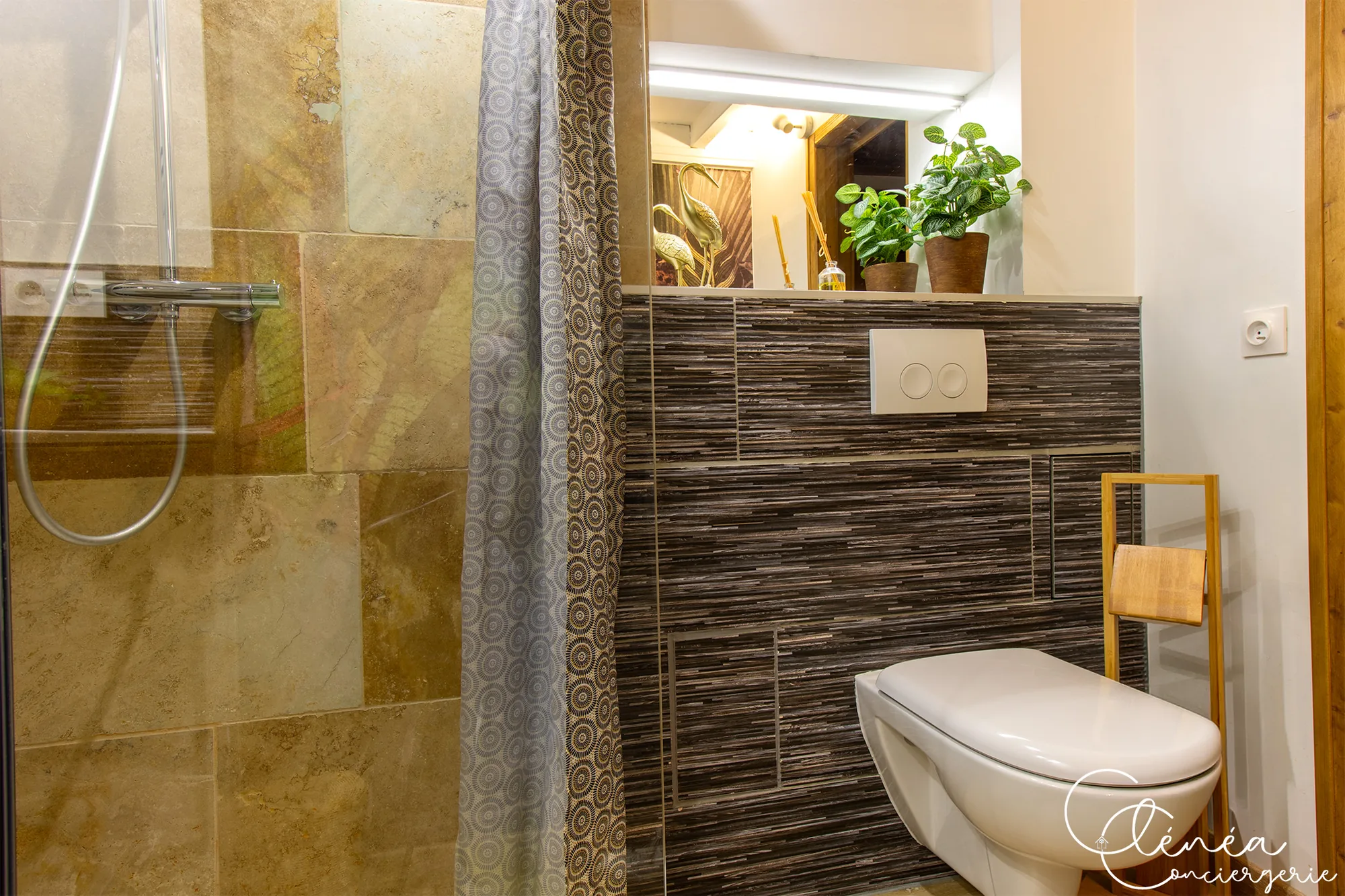 The Nest — Douche avec paroi vitrée et carrelage beige, à côté d’un WC suspendu et d’un porte-serviettes en bois.