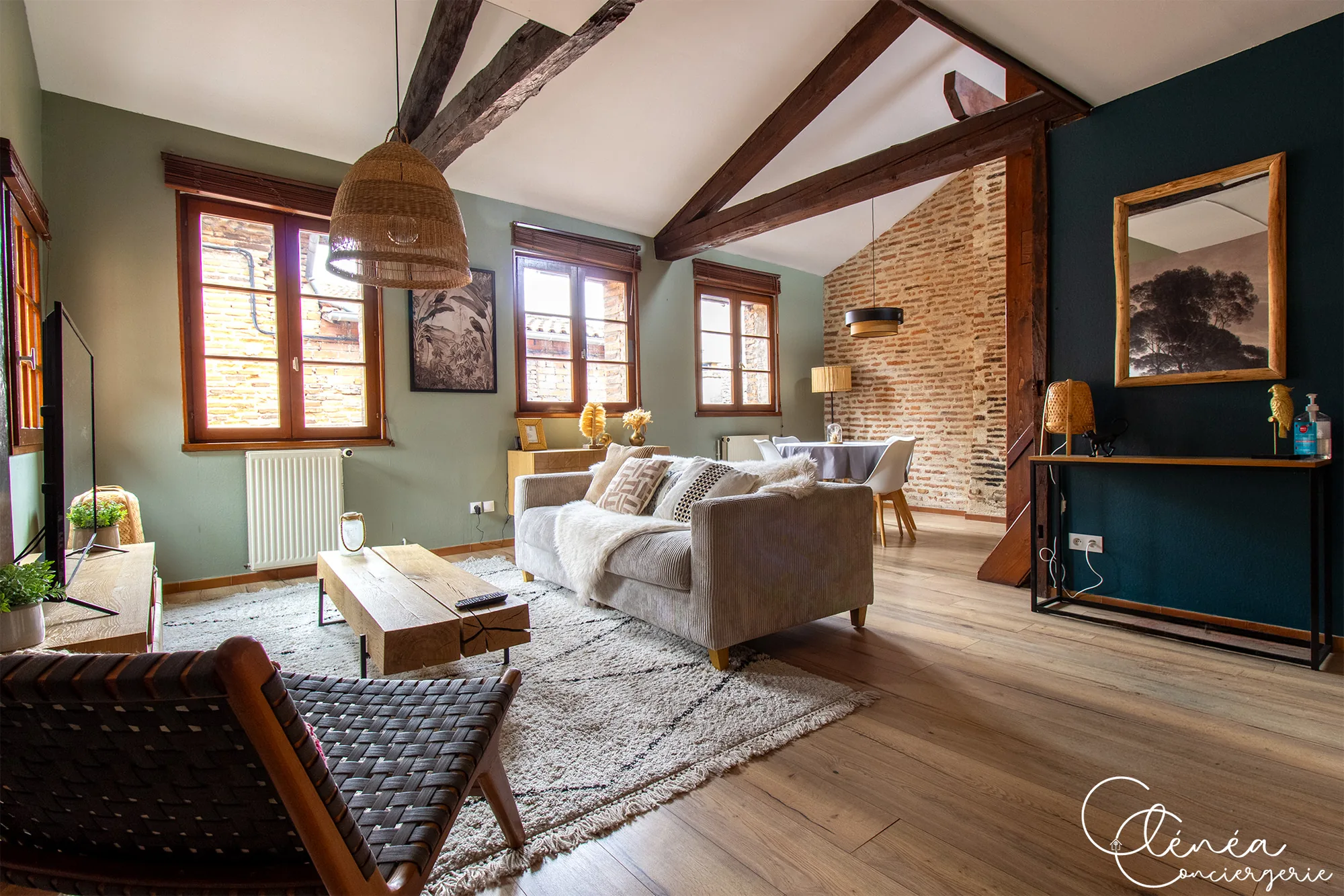 The Nest — Vue du salon vers un pan de mur bleu foncé avec console en bois et miroir, le coin repas au fond contre le mur de briques.