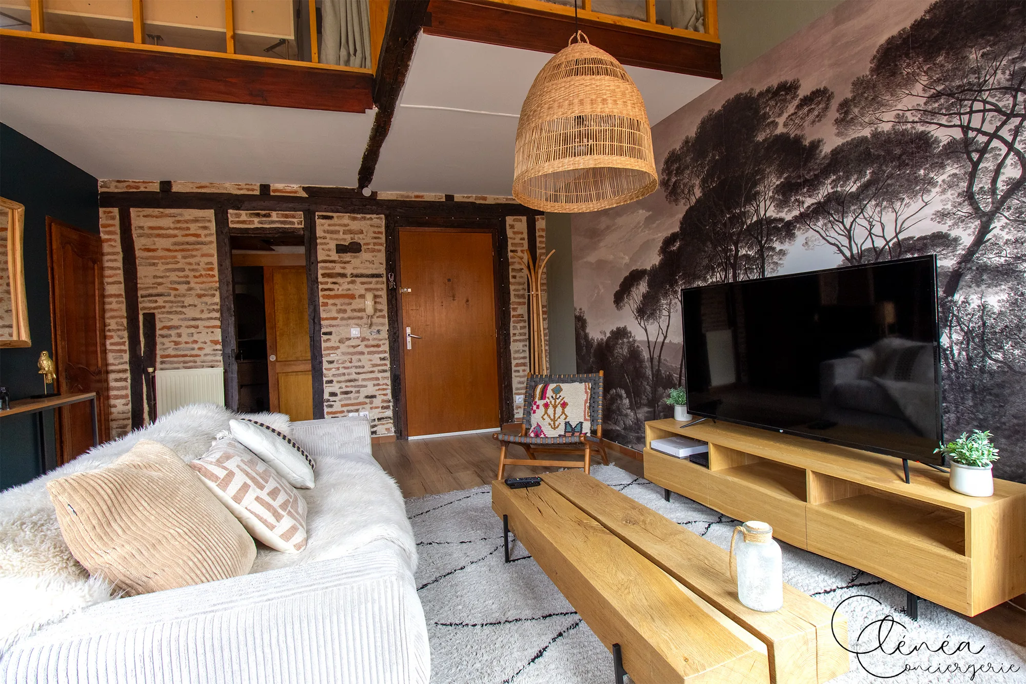 The Nest — Salon avec grande télévision devant une fresque murale d’arbres, porte d’entrée en bois et mezzanine surplombant la pièce.