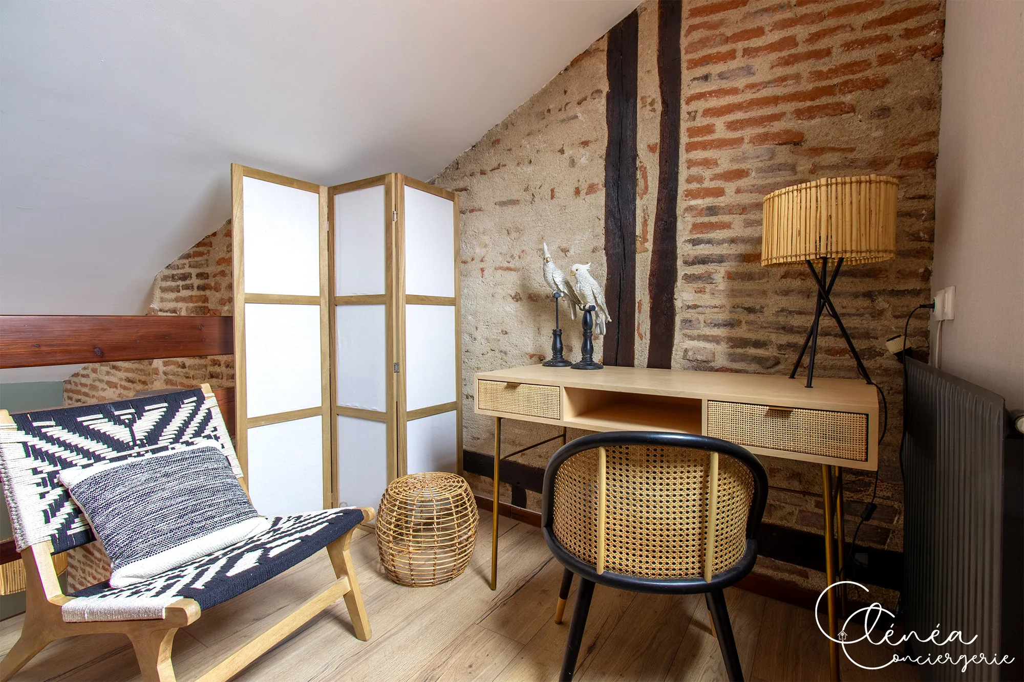 The Nest — Bureau en bois avec chaise cannée et lampe, devant un mur de briques et colombages, à côté d’un fauteuil et d’un paravent.