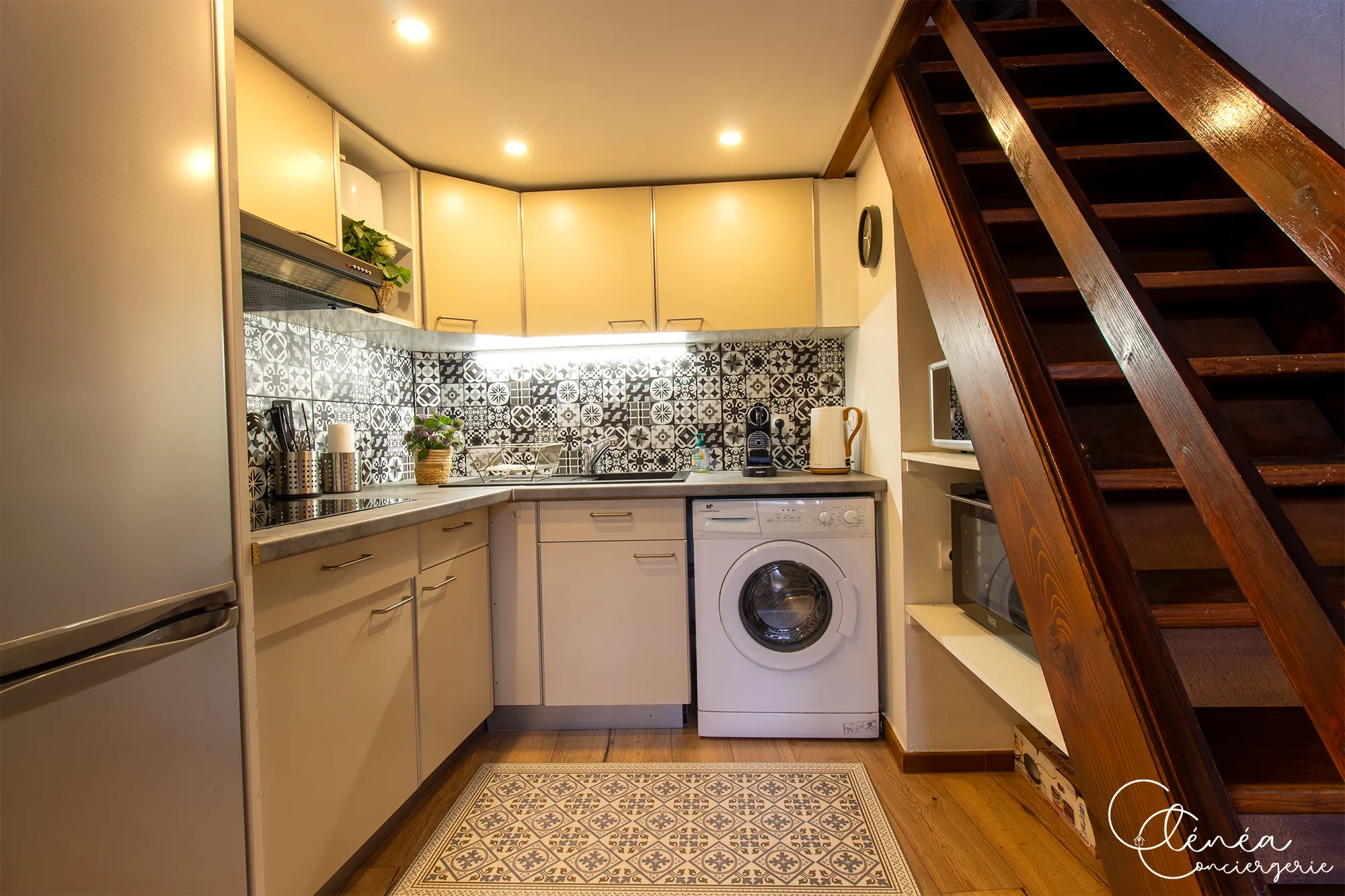 The Nest — Cuisine vue d’ensemble avec réfrigérateur, plaques, lave-linge et micro-ondes rangé sous l’escalier en bois.