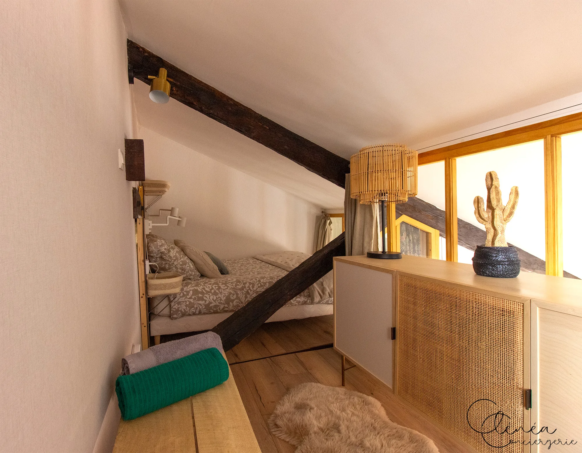The Nest — Chambre de la mezzanine avec lit double sous les poutres, banc portant des serviettes et meuble de rangement en cannage.