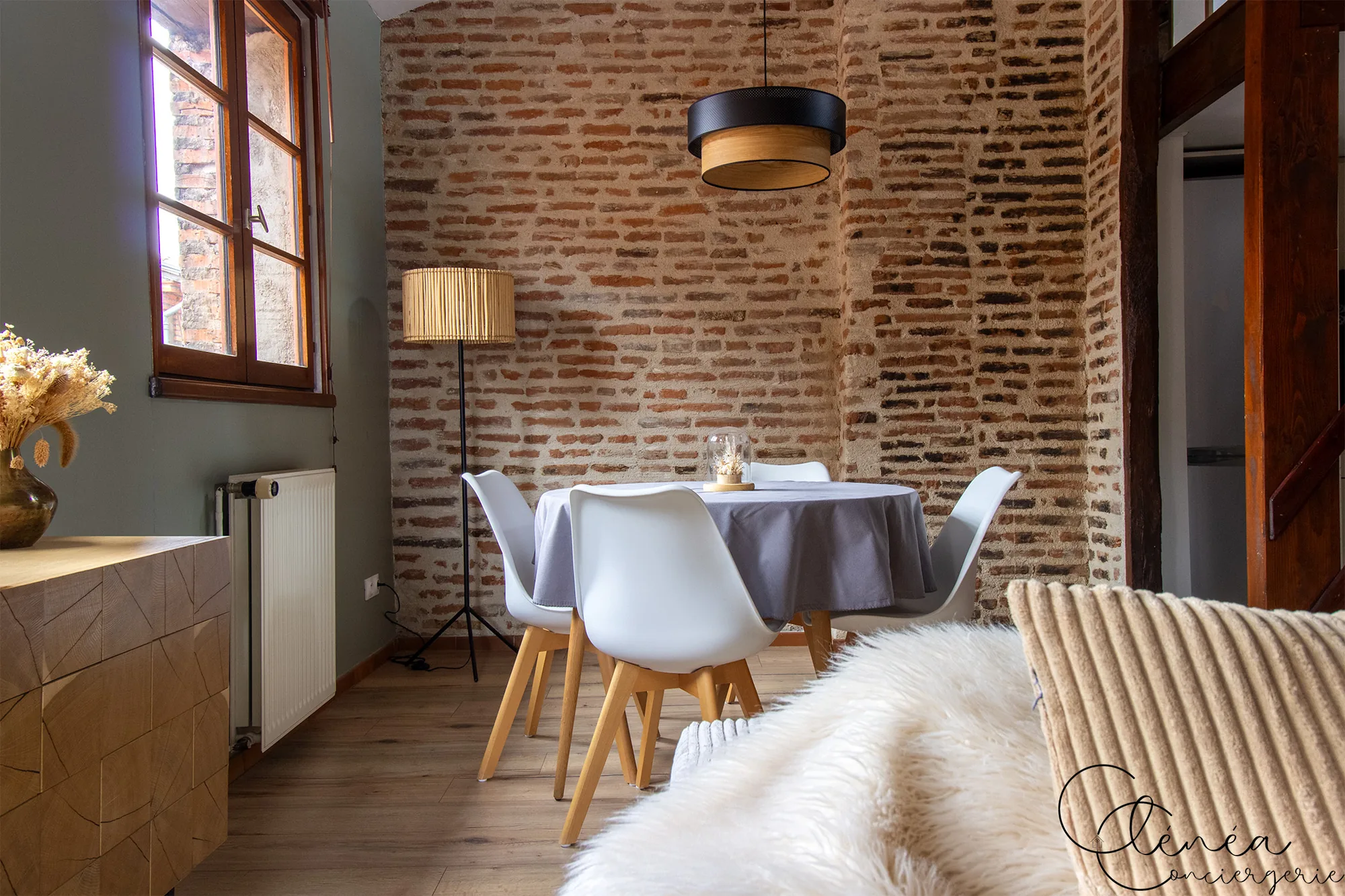 The Nest — Table ronde nappée de gris entourée de quatre chaises blanches, devant un mur de briques apparentes.