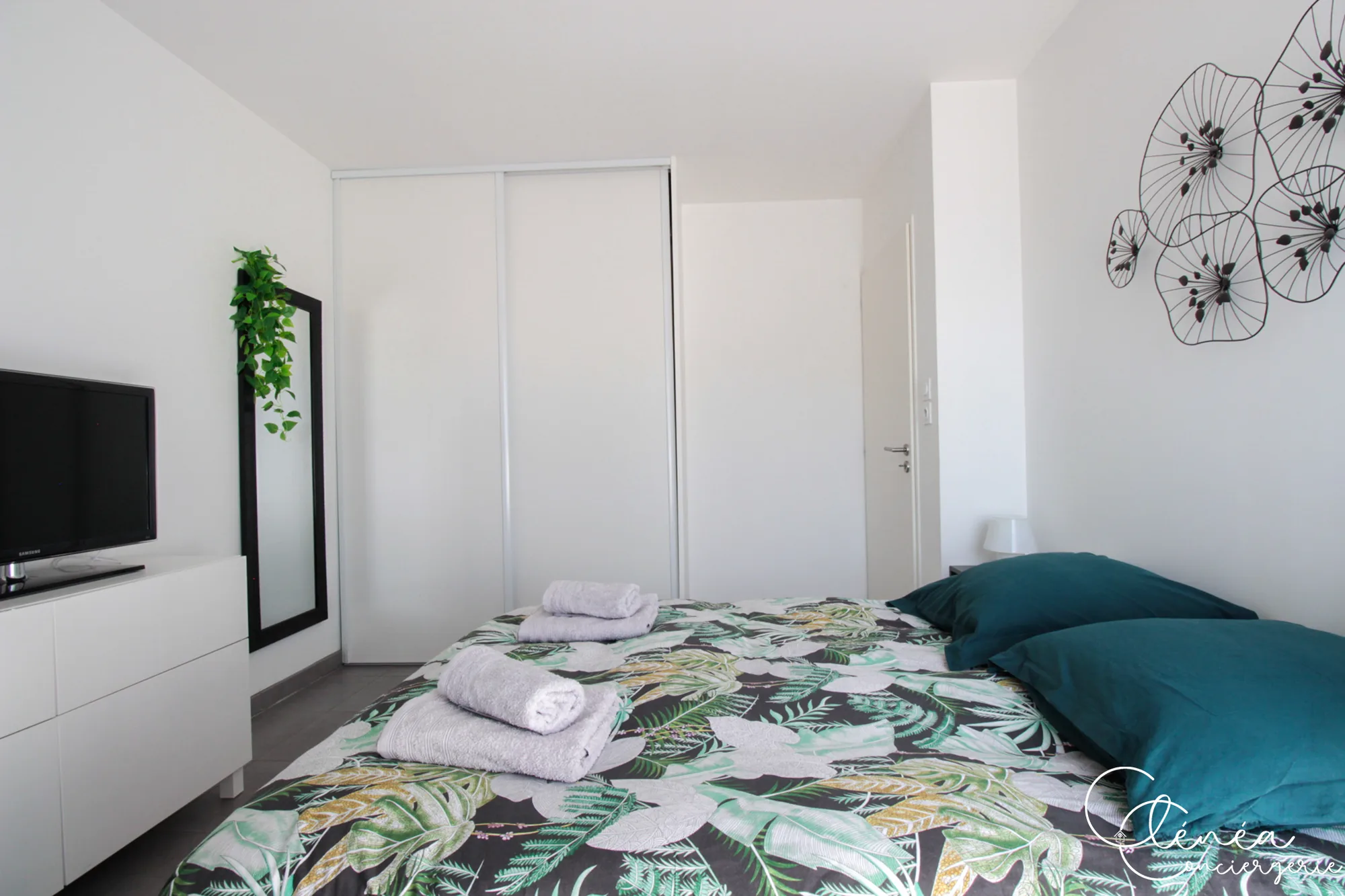 Urban Jungle — Chambre avec linge de lit à motif de feuillage, télévision posée sur une commode blanche et grand placard à portes coulissantes.