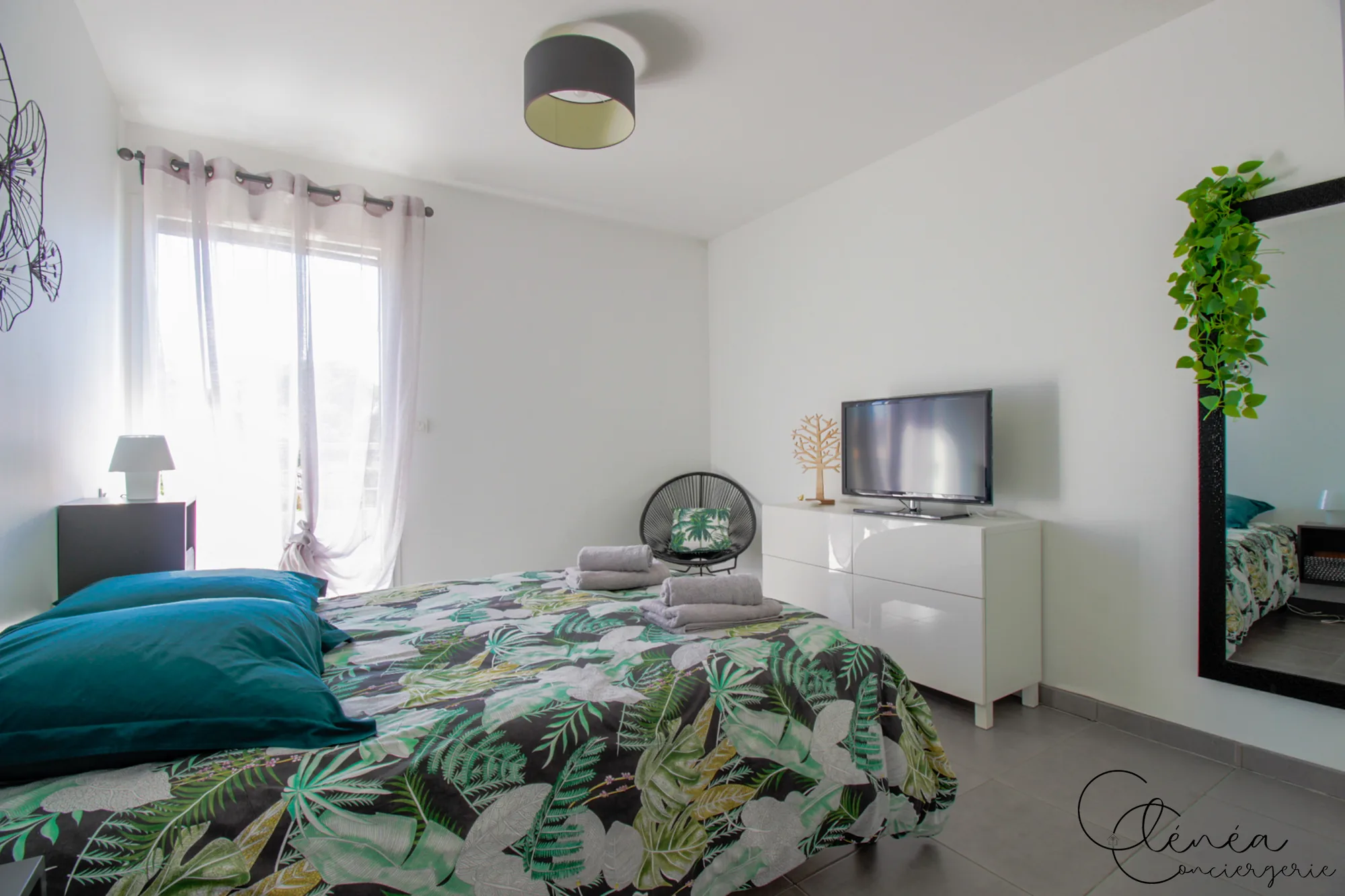 Urban Jungle — Chambre au décor végétal avec lit double, fenêtre habillée d’un voilage, fauteuil rond noir et télévision sur la commode.