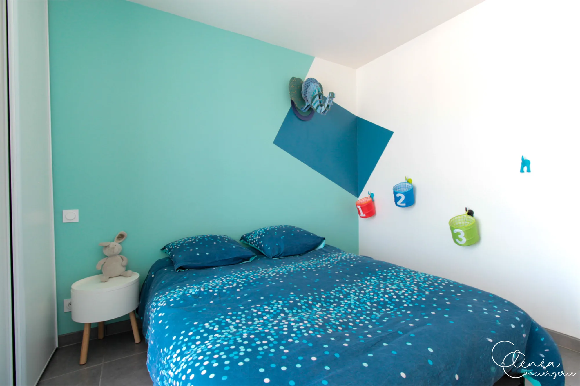 Urban Jungle — Chambre aux murs turquoise avec lit double, table de chevet ronde et paniers de rangement numérotés fixés au mur.