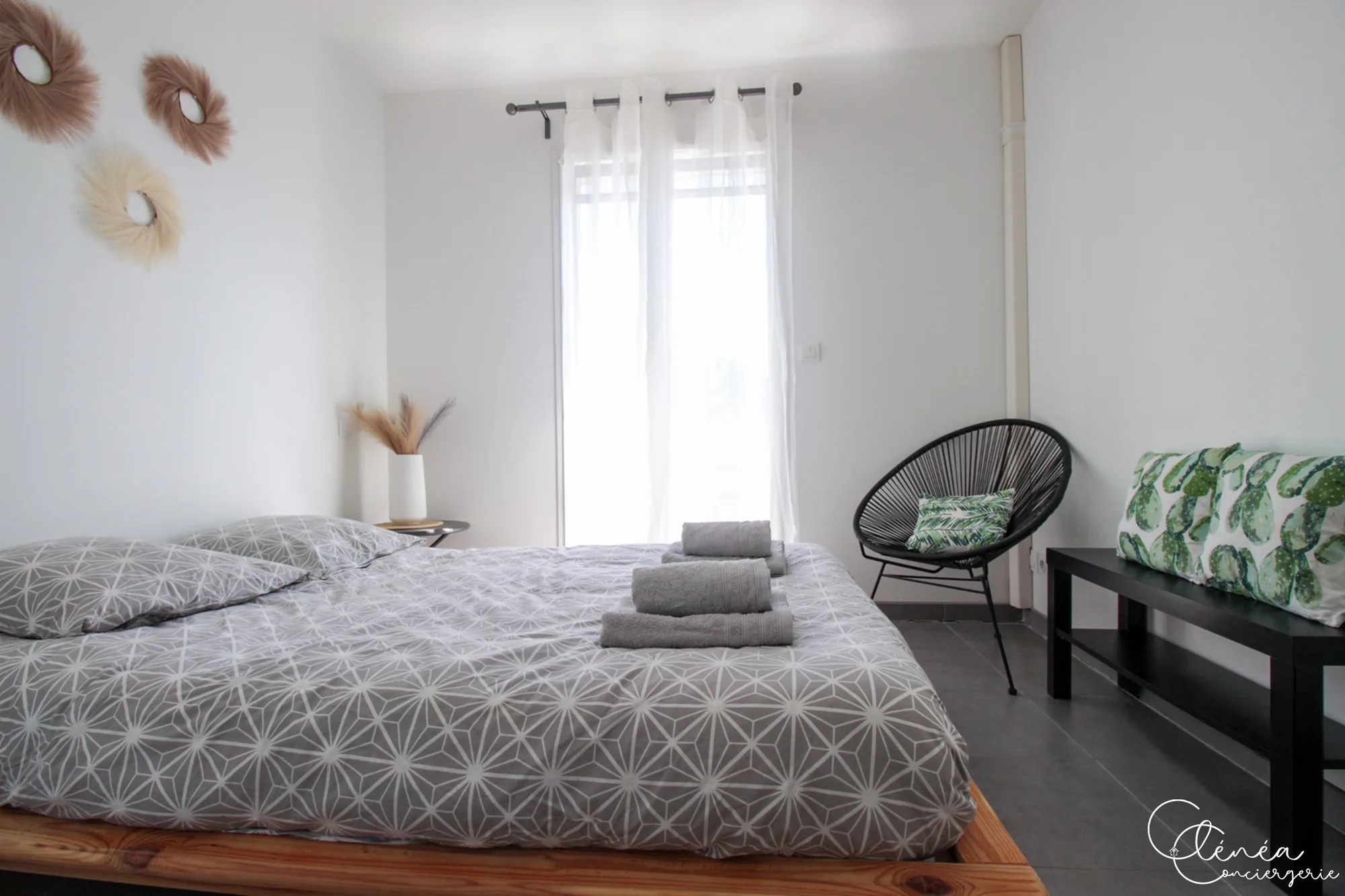 Urban Jungle — Chambre claire avec lit double sur cadre en bois, housse de couette grise à motifs et fauteuil rond noir devant la fenêtre.