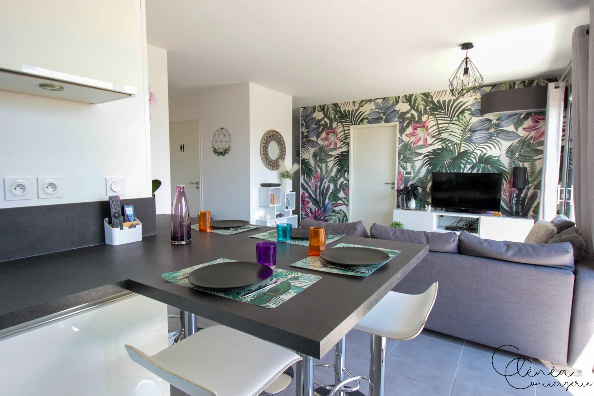Urban Jungle — Table haute dressée pour le repas, ouverte sur le salon et son mur habillé d’un papier peint tropical.