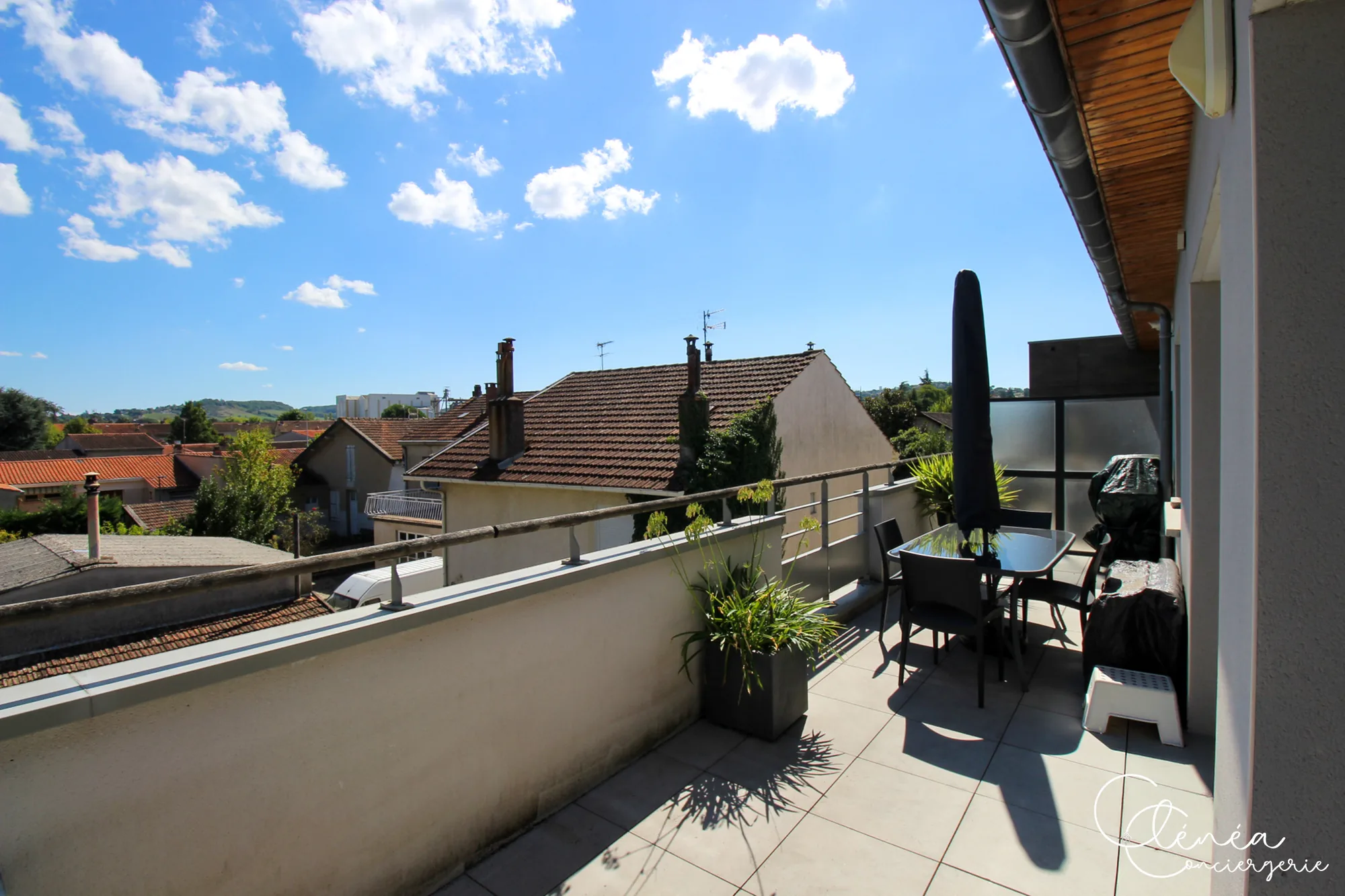 Urban Jungle — Terrasse avec petite table et quatre chaises, grand parasol fermé et barbecue protégé par une housse, face aux toits.