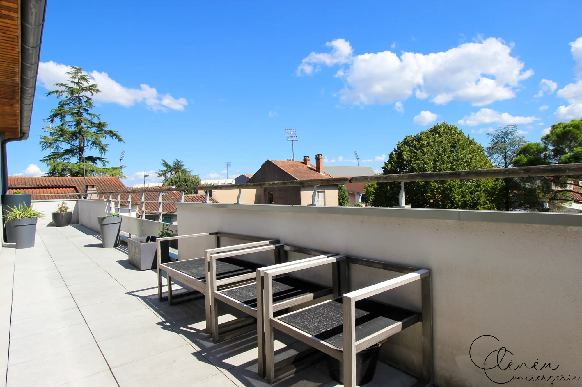 Urban Jungle — Trois fauteuils en bois grisé alignés le long du muret de la terrasse, avec vue sur les toits et les arbres.