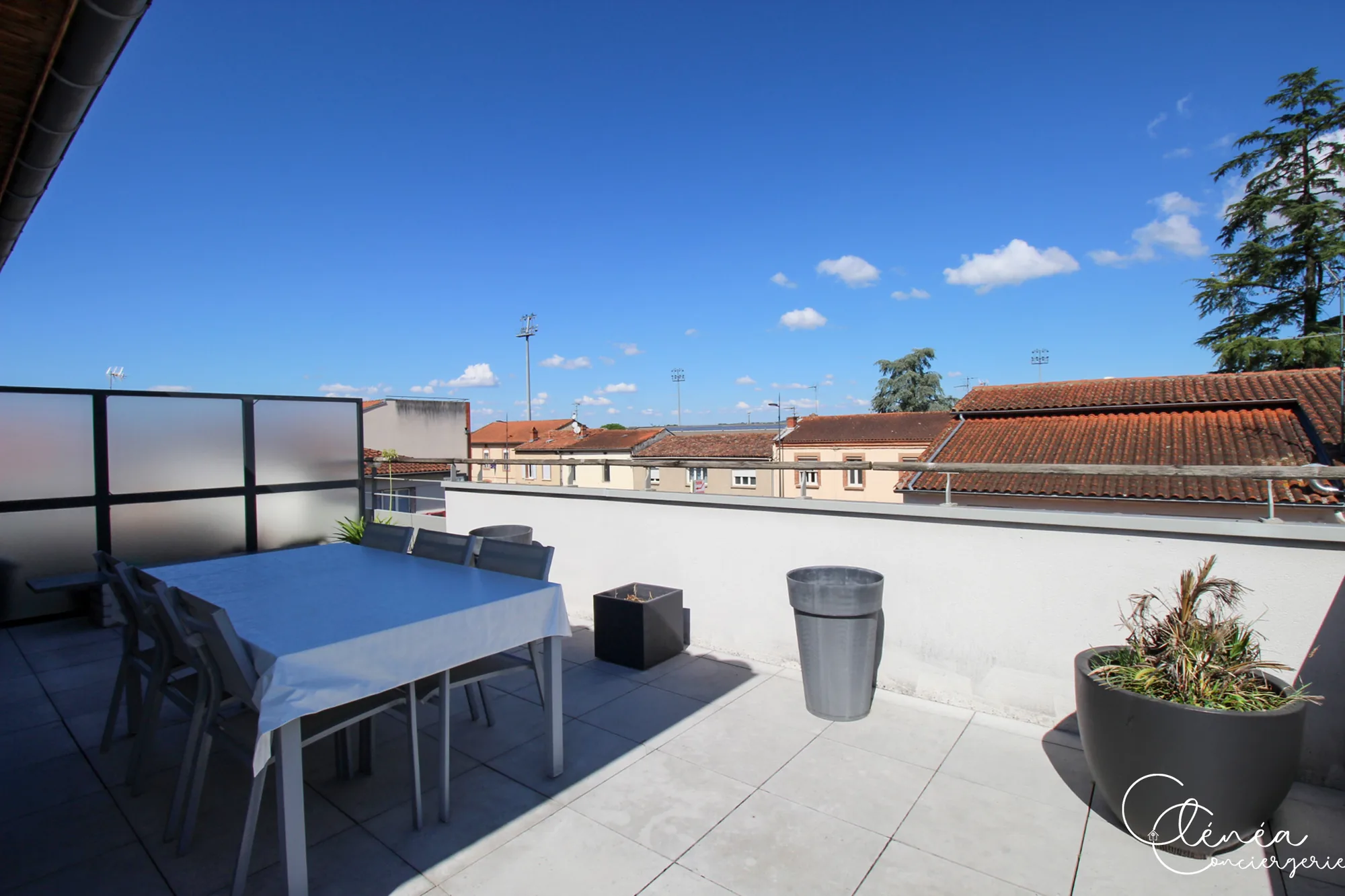 Urban Jungle — Table de terrasse recouverte d’une nappe et entourée de chaises, devant un panorama de toits et de cèdres.