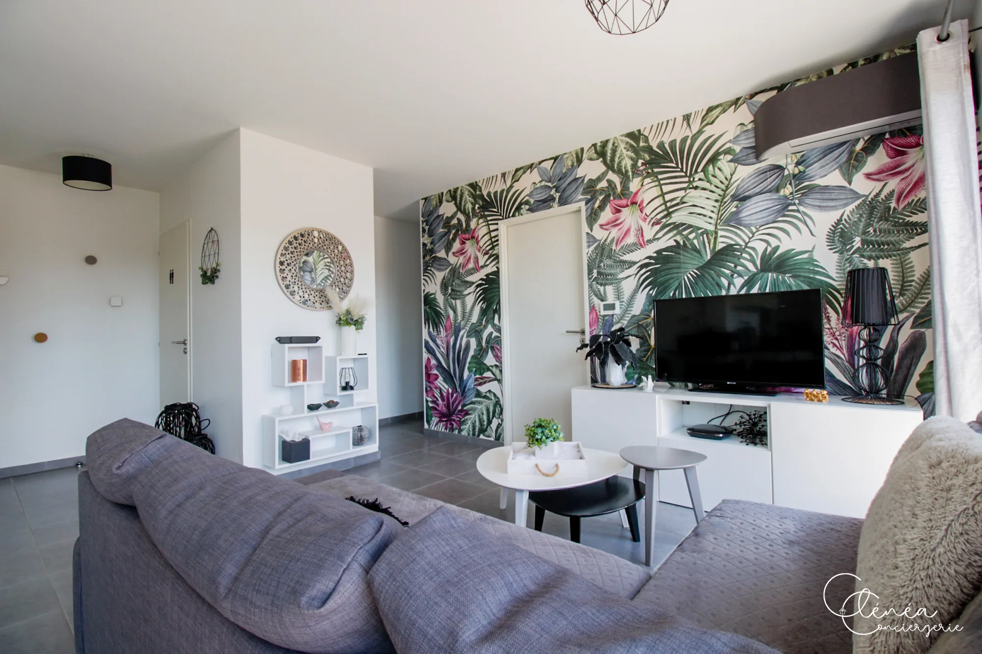 Urban Jungle — Salon vu depuis le canapé, avec étagères décoratives, miroir rond et mur au papier peint tropical.