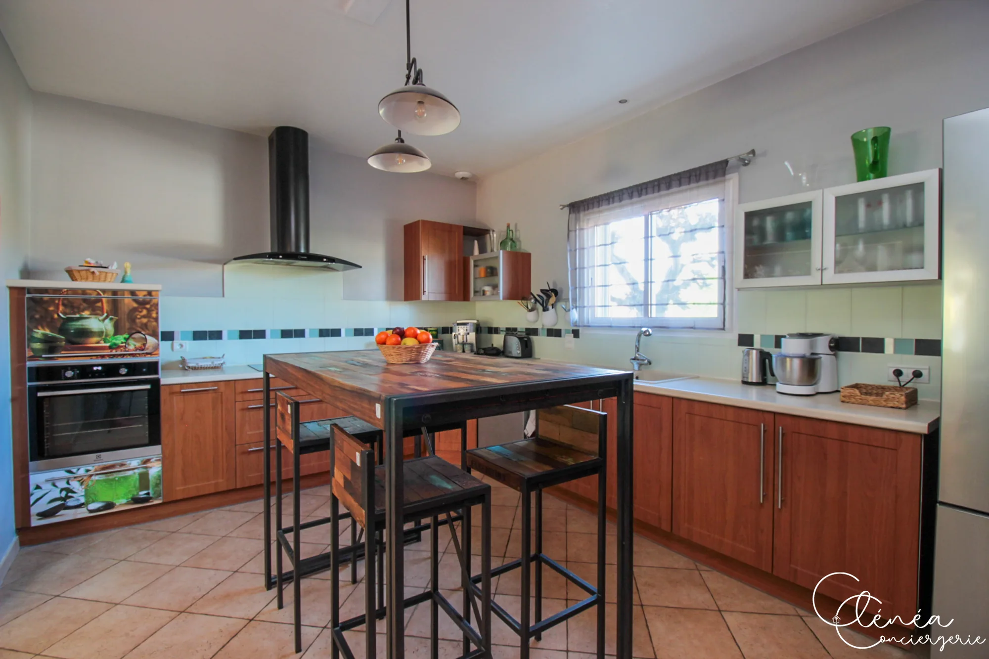Villa Chanoine — La cuisine équipée avec sa table haute en bois et ses tabourets, son four, sa hotte et ses plaques de cuisson.